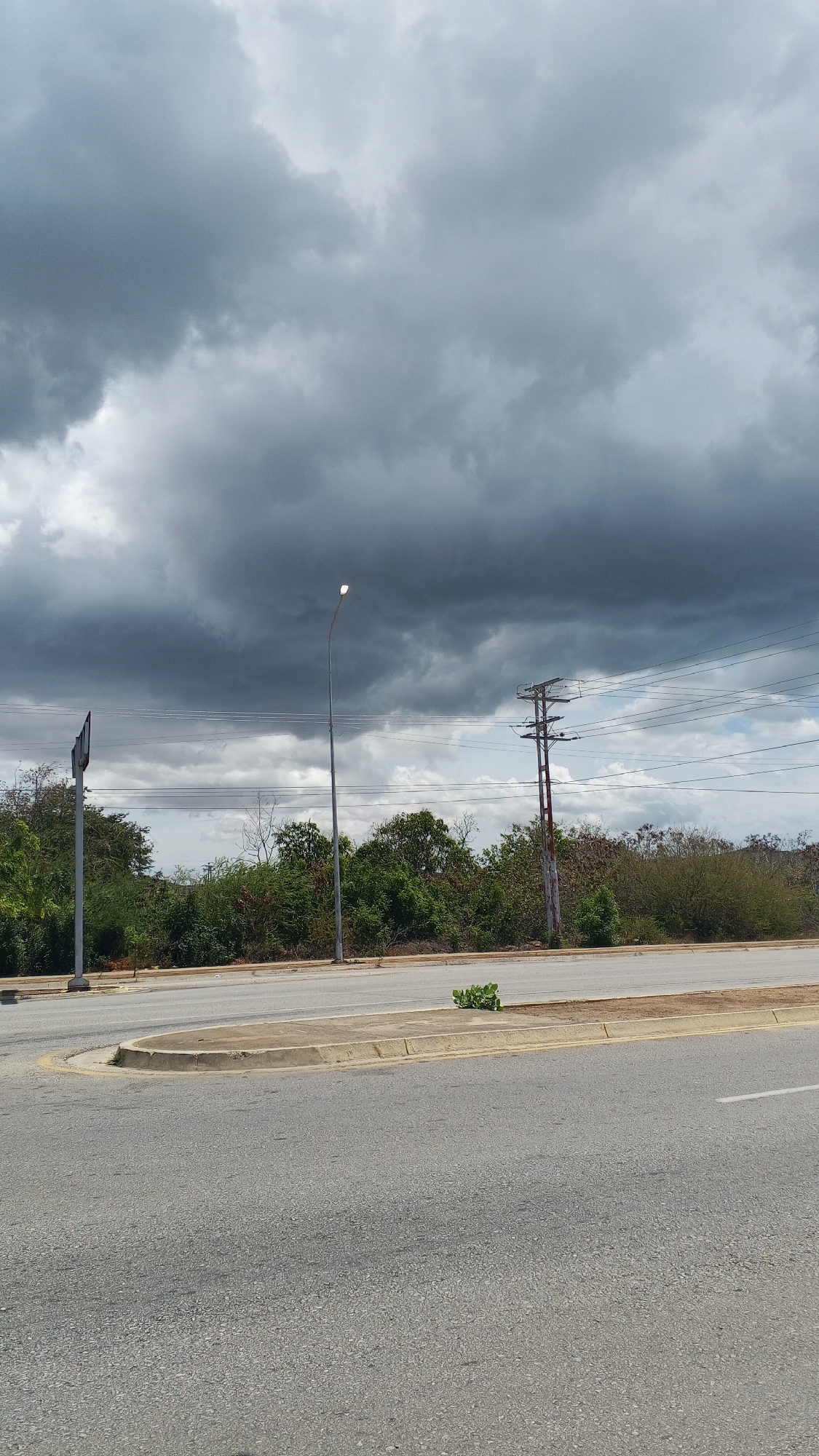 The image depicts an urban scene in Unknown, Unknown, characterized by an overcast sky with dark, ominous clouds dominating the upper portion of the frame. The lower portion is dominated by a grey asphalt road with a white lane marker and a median or curb.
In the background, there is a line of green vegetation, interspersed with utility poles and power lines stretching across the horizon. There are two visible poles, one a standard street light with its bulb illuminated, and another, taller utility pole with crossbeams. A sign can be seen in the distance, next to the streetlight.
The overall impression is one of a potentially stormy day in an urban or suburban environment, with a somewhat subdued atmosphere due to the heavy cloud cover. There are no people or animals directly visible in the frame.