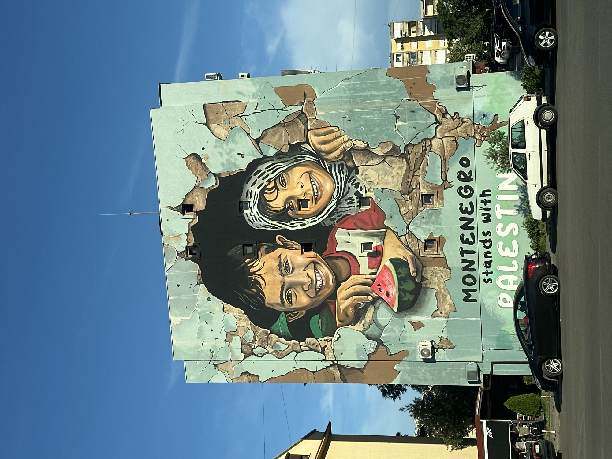 A large mural depicting two smiling children covers the side of a building under a clear blue sky. The mural uses a trompe l'oeil effect, making it appear as if the children are breaking through a cracked, pale blue wall. One child, wearing a white and black keffiyeh, is positioned at the top left, reaching out with an open hand. The other child, at the bottom right, holds a slice of watermelon and also smiles broadly.
To the right of the children, vertically oriented text reads "MONTENEGRO stands with PALESTIN". The last two letters of "PALESTINE" are obscured or cut off. The building's facade is also visible, with small windows incorporated into the mural design.
Several cars are parked along the street to the right of the building, including a white car and a black car. Other buildings are visible in the background, suggesting an urban setting. The overall scene is brightly lit by what appears to be daytime sunlight. The location is Podgorica, Montenegro.