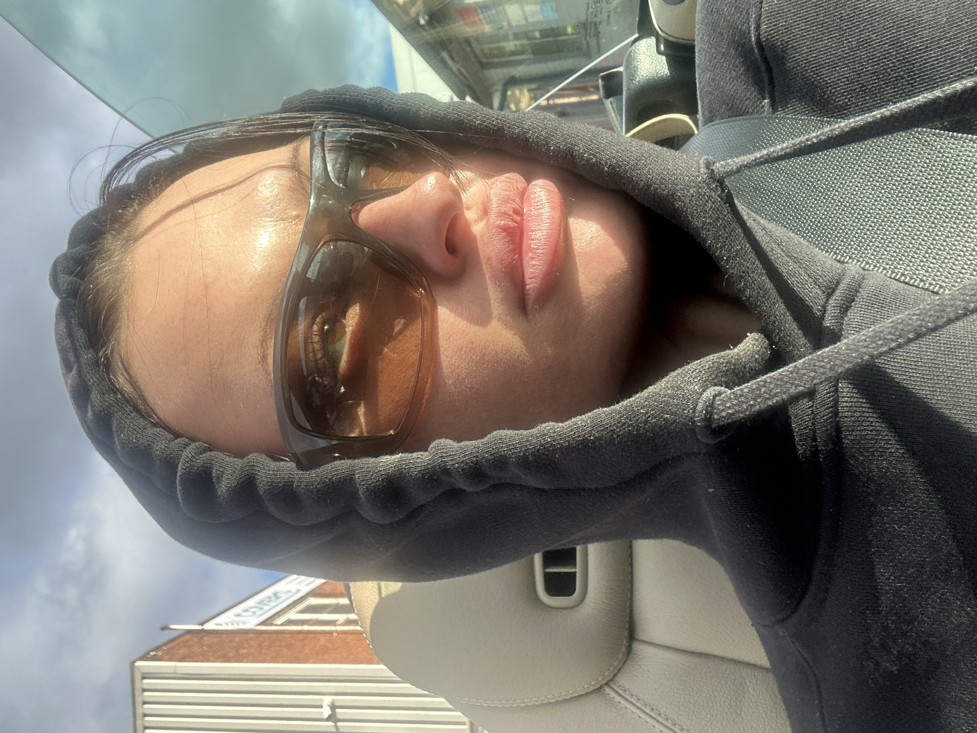 The image displays a female individual situated within a vehicle on Longfield Road, Tunbridge Wells, United Kingdom. The subject, visible from the chest upwards, wears a dark-colored hooded sweatshirt with the hood up, and dark brown rectangular sunglasses. Her lips are slightly pursed, and dark hair strands are visible near her forehead. A seatbelt traverses diagonally across her chest. She is seated in a vehicle with a light beige/tan leather seat headrest visible behind her. The right side of her face is brightly illuminated by direct sunlight.
In the background, visible through the vehicle's windows, are commercial buildings. To the left, a structure with a red brick facade stands next to a building featuring light-colored vertical corrugated metal siding. A sign partially displaying the word "CORK" is visible between these two structures, confirming the location as Longfield Road near Cork Storage Solutions. The sky overhead shows scattered clouds with patches of blue. The scene is illuminated by strong, direct sunlight, indicating daytime.