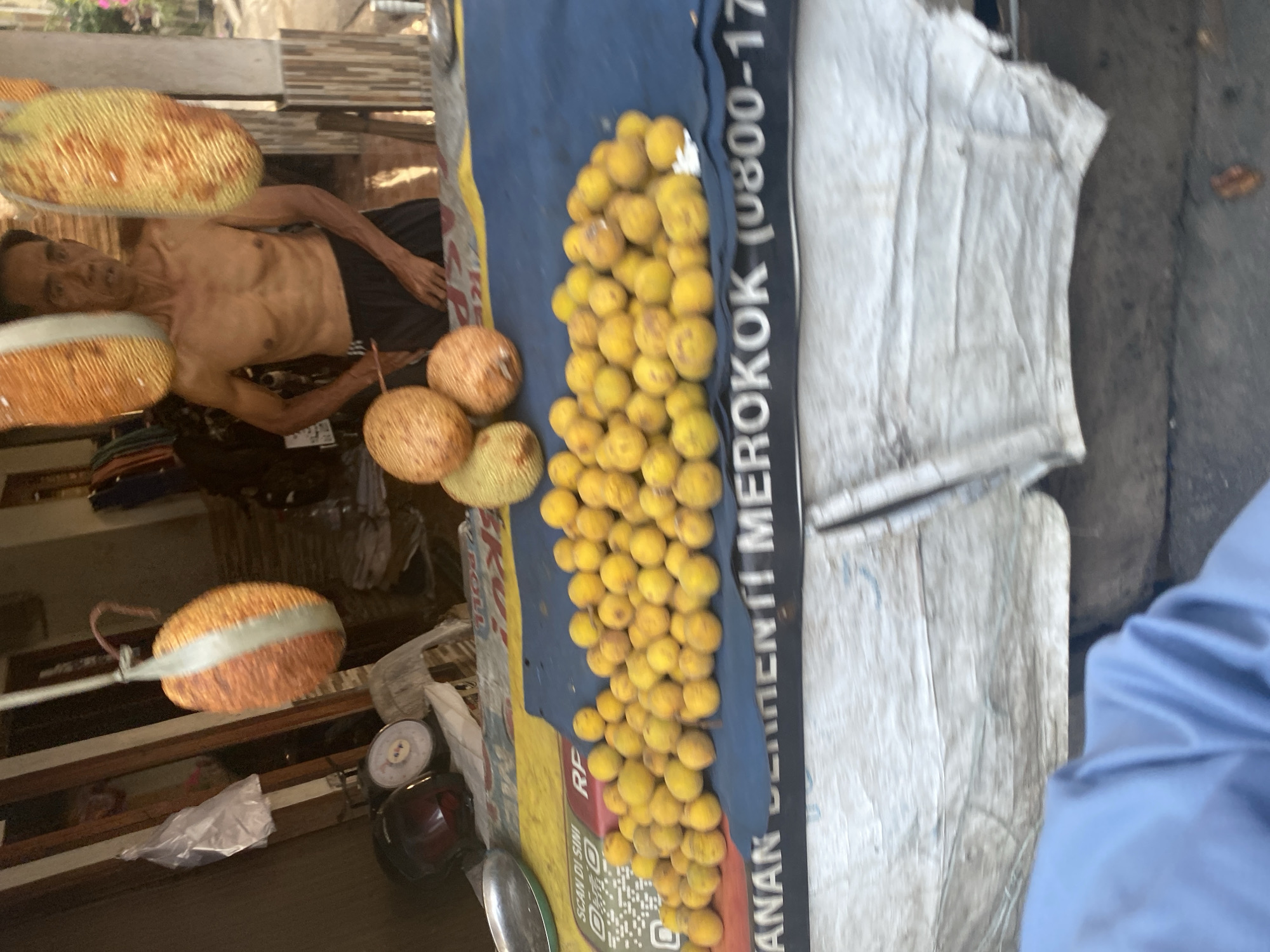 A man is standing behind a market stall in Depok, Indonesia, selling fruit. He is shirtless and wearing black shorts. He is surrounded by jackfruit and a pile of small, yellow fruits, possibly citrus. A scale and a helmet are visible on the stall. A blue tarp is spread out on the stall, covered with the fruit. In the background, there are buildings and greenery. The image is taken from a low angle, suggesting the photographer is standing.