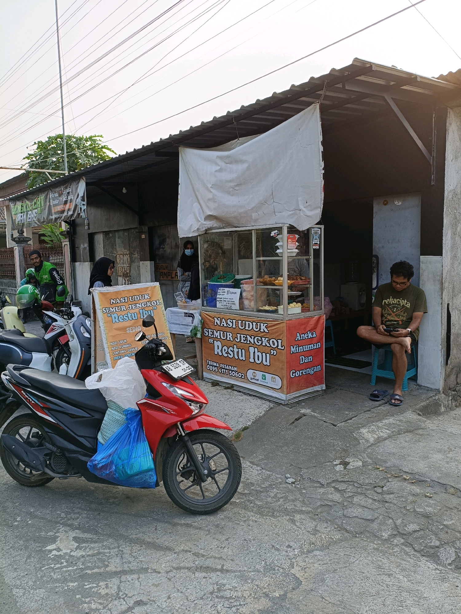 In Maguwoharjo, Indonesia, a street-side food stall displays "Nasi Uduk Semur Jengkol Restu Ibu" alongside "Aneka Minuman Dan Gorengan." A red motorcycle is parked in the foreground, laden with blue plastic bags and white fabric. To the left, a man in a green Gojek jacket and helmet stands near a white scooter, while a woman in a hijab stands beside the food stall. To the right, a man in a dark green t-shirt sits on a blue stool, engrossed in his phone. The stall has a glass display case showcasing various food items. The environment appears to be a humble urban setting with a corrugated metal roof extending over the stall and a concrete ground surface. Overhead, a dense network of power lines stretches across the sky.