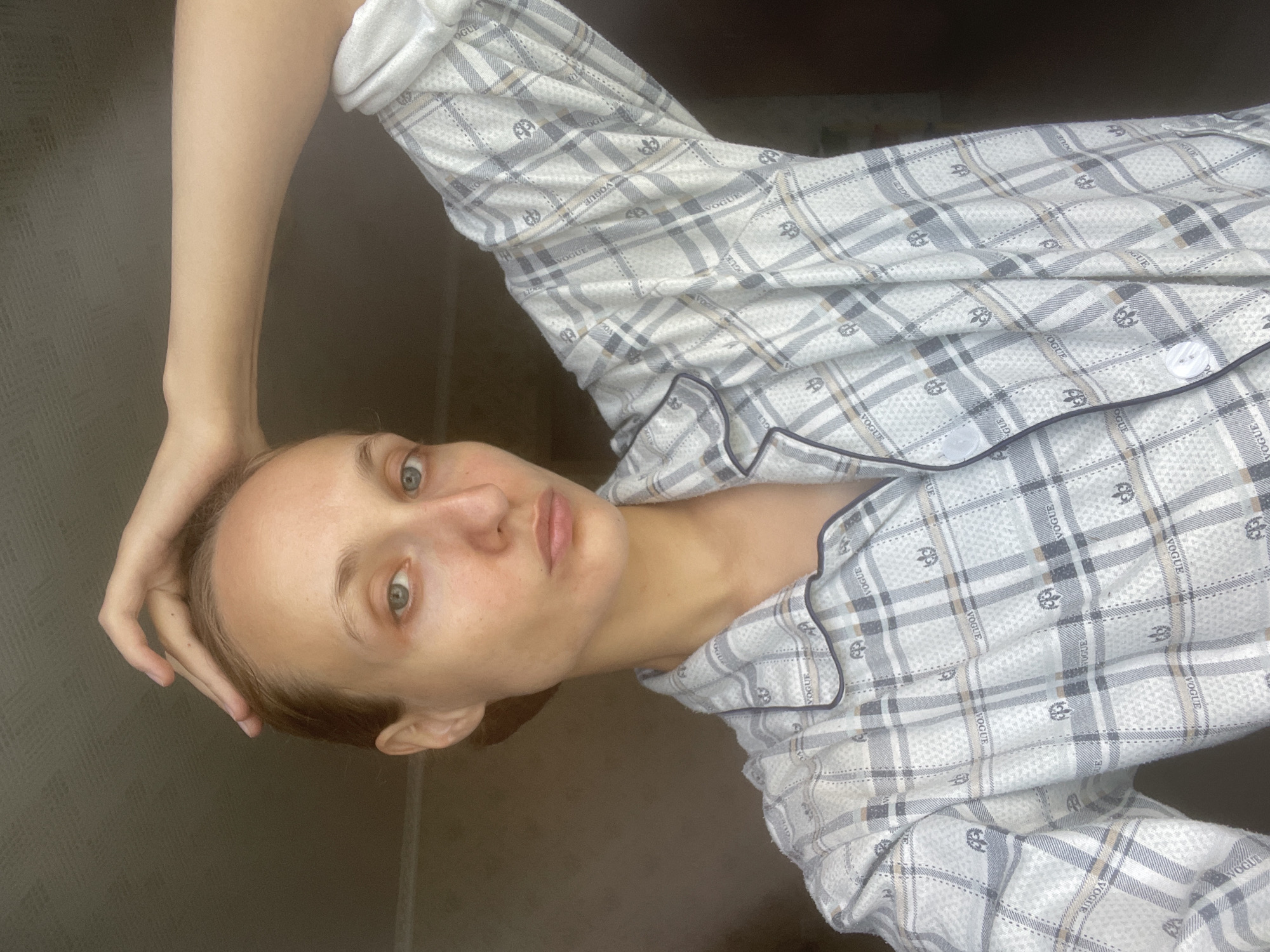 A woman is shown from the chest up, with her right arm raised and her hand resting on her head. She is wearing pajamas with a plaid pattern. Her hair is pulled back, and she is looking directly at the camera. The background is plain and appears to be indoors, with no discernible city elements.