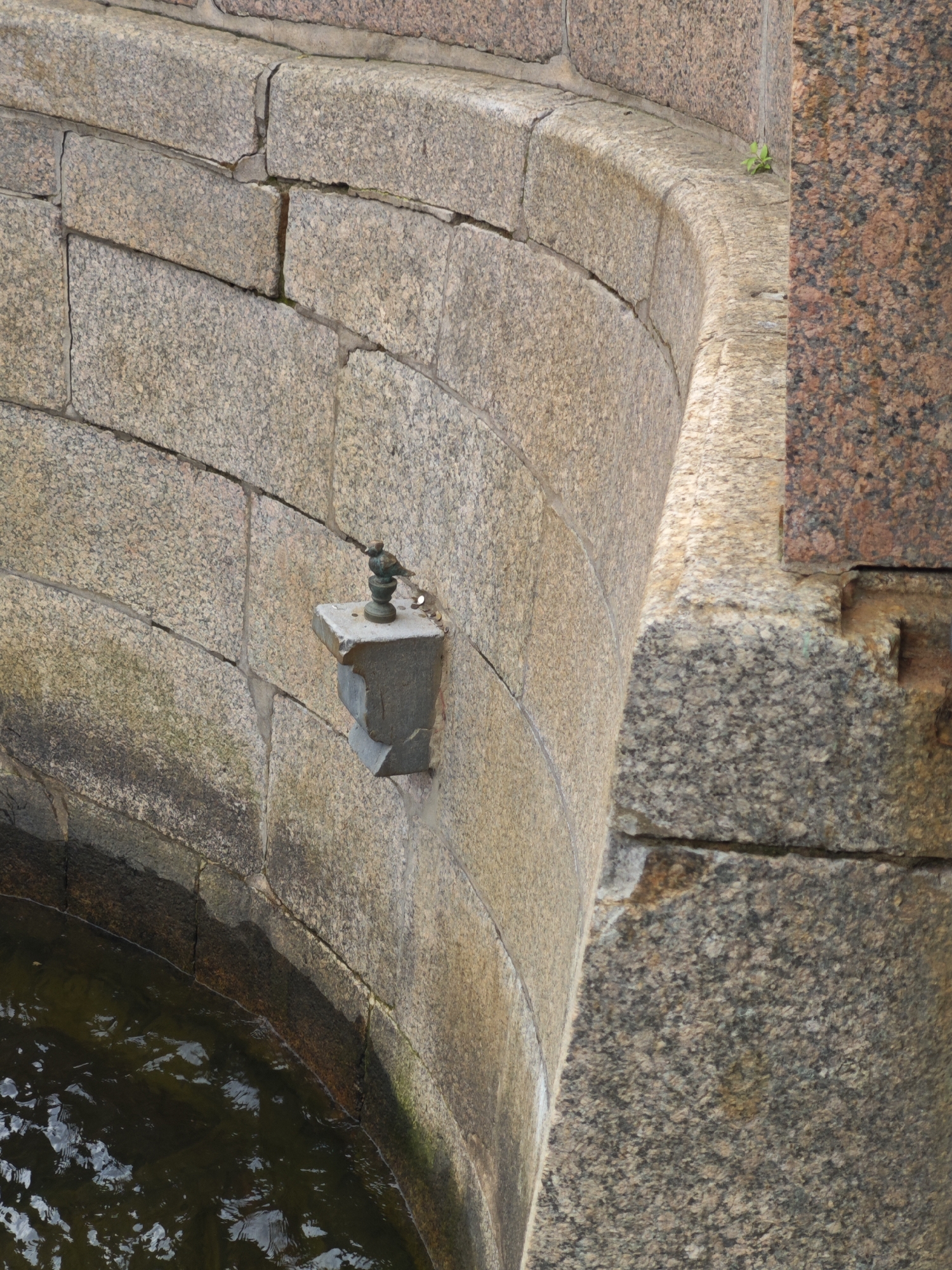 The image shows a close-up view of the granite embankment of a canal or river in Saint Petersburg, Russia. The embankment is constructed from large, rough-hewn granite blocks, some of which show signs of wear and discoloration, with patches of moss and algae in places. Water is visible at the bottom of the frame, reflecting the overcast sky. A small, decorative bronze bird sculpture is mounted on a stone bracket attached to the embankment. The bird is facing to the right, and the bracket appears to be slightly chipped. Coins are scattered on the top surface of the bracket. The overall scene depicts a detail of the city's historic infrastructure, likely along one of its many waterways.