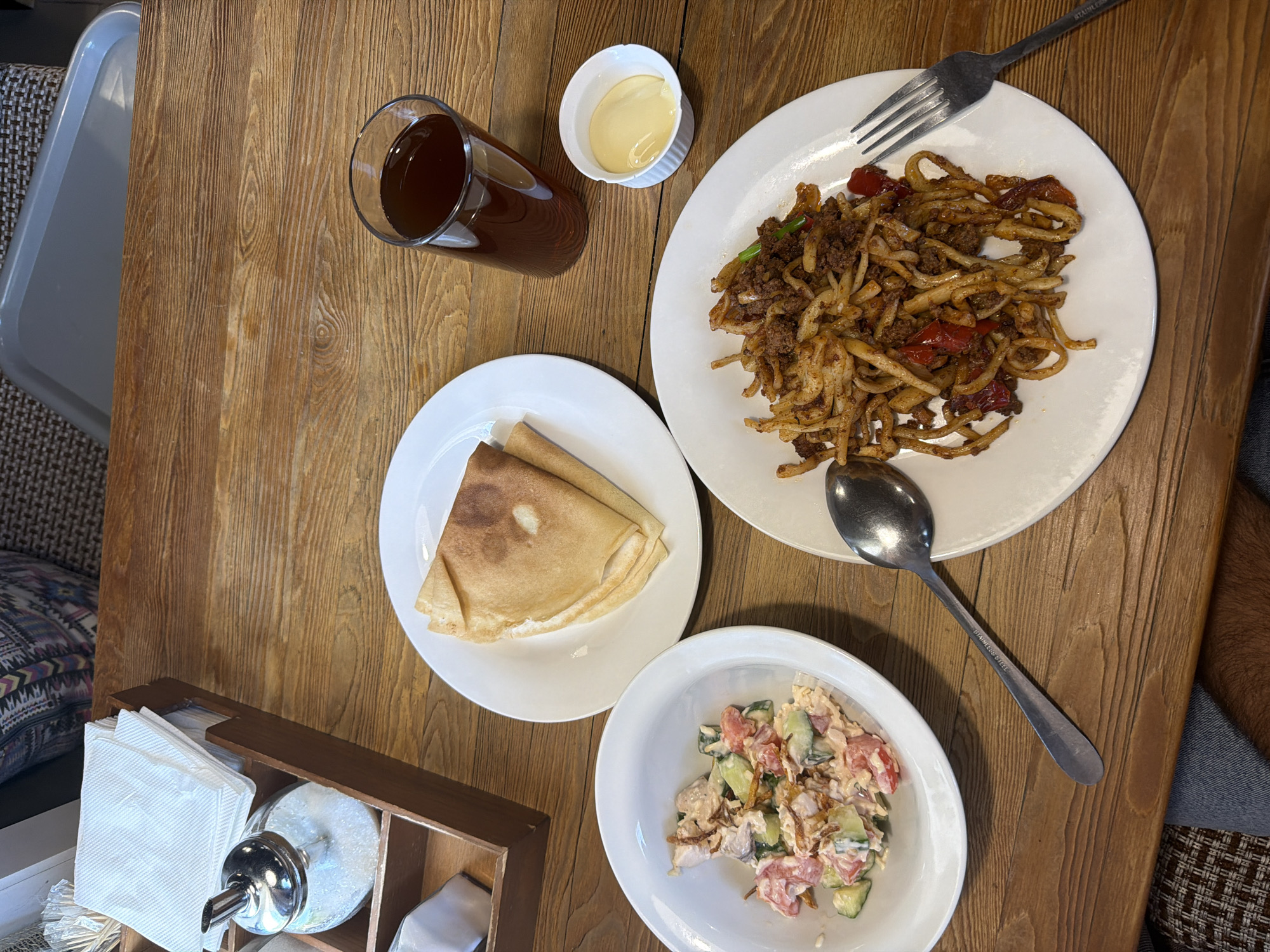 An overhead shot captures a meal laid out on a light brown wooden table, likely in a cafe or restaurant in Krasnoyarsk, Russia. The primary dish, a plate of noodles mixed with ground meat, red bell peppers, and possibly other vegetables, is positioned to the right, with a metal fork resting above it. The fork handle has "STAINLESS" visible. Below this, a white bowl contains a salad of diced tomatoes, cucumbers, and what appears to be shredded chicken or a similar protein, tossed in a creamy dressing. A metal spoon lies between the main dish and the salad.
To the left, a white plate holds two folded blini or crepes. In the upper left corner, a tall glass contains a dark-colored drink, possibly tea or kvass, next to a small white ribbed bowl holding a light yellow sauce or condiment. A wooden caddy, containing white paper napkins and a glass sugar dispenser, is visible in the lower-left foreground. Portions of a gray tray and a patterned cushion are visible on the left side, indicating seating. A person's arm or leg is partially visible on the far right, suggesting someone is seated at the table. The lighting indicates it is daytime.