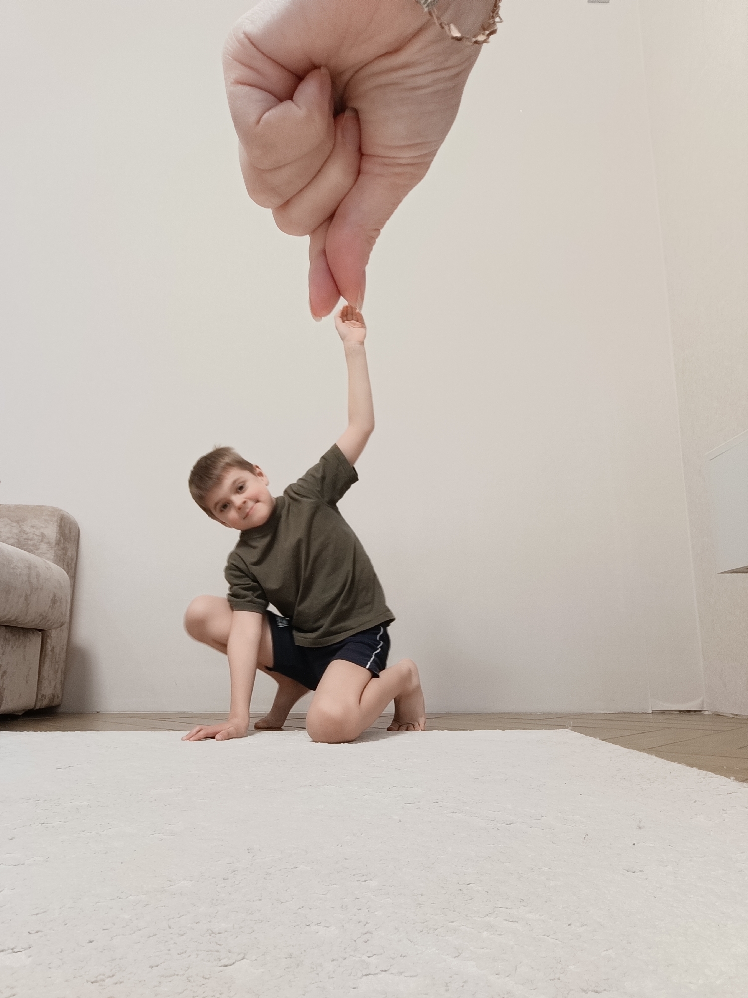 A young boy with short brown hair is kneeling on a white rug in a room with light beige walls. He is wearing a dark green T-shirt and dark blue shorts with white stripes. His left arm is raised, and his hand is being pinched by the index finger and thumb of a much larger hand that appears to be suspended in the air above him. The larger hand has pale skin and is adorned with a gold-colored bracelet or watch. Part of a light-colored couch is visible in the background to the left. A light-colored rectangular object is on the wall behind the boy to the right. The location of the photograph is unknown, and there is no information that shows this image took place in the city of Chaikovsky, Russia.