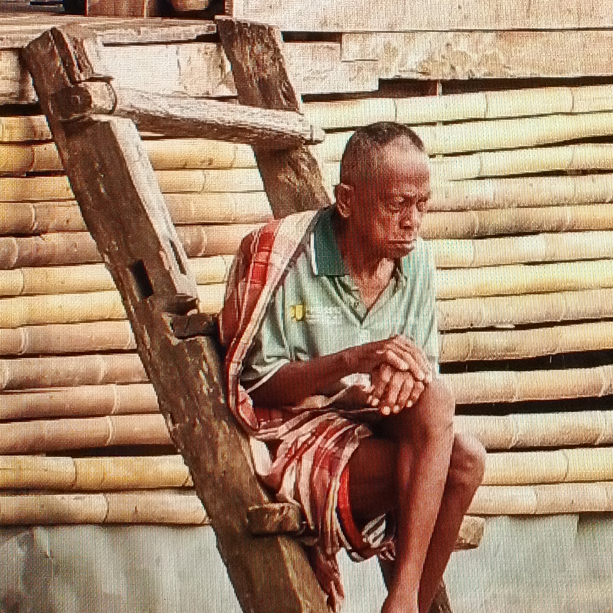 A man is sitting on a wooden structure. He is wearing a light green shirt with a V-neck and a red and white checkered sarong or cloth draped over his shoulders and around his waist. His hands are clasped in front of him. The background consists of a wall made of horizontal wooden planks. The overall scene appears to be outdoors, possibly in a rural or traditional setting in Ende, Indonesia. The lighting suggests it is daytime.