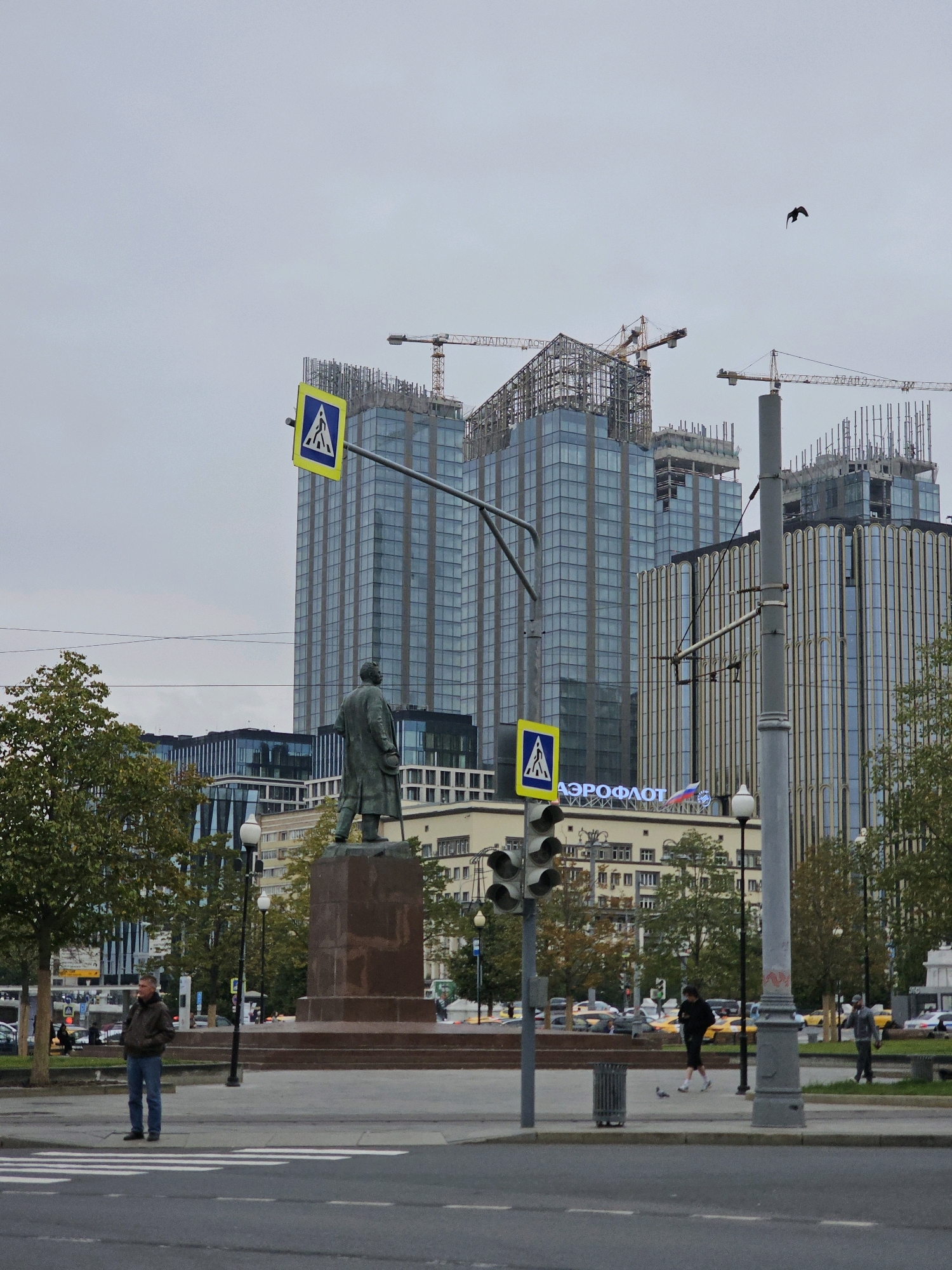 This image depicts a street scene in Moscow, Russia, likely near the Aeroflot building, identified by the prominent signage. In the foreground, a pedestrian crossing with white stripes is visible on a grey asphalt road. A man in a brown jacket and blue jeans stands on the sidewalk to the left, facing away from the camera. To the right, a traffic light pole with a pedestrian crossing sign is present.
Behind the pedestrian area, a large bronze statue of a man on a stone pedestal stands prominently. The statue is the focal point of the midground, set against a backdrop of modern architecture.
In the background, several tall, glass-covered buildings are under construction, indicated by visible cranes and scaffolding. One of these buildings displays the "AEROFLOT" logo in large letters, with a Russian flag above it. The sky is overcast and grey. Trees with green foliage line the sides of the pedestrian area, suggesting it might be autumn or late summer.
Several people are visible walking on the sidewalks and crossing the street in the distance. Cars and yellow taxis are present on the road. A lone bird is captured in flight against the grey sky. The overall scene conveys an urban environment with a mix of historical elements (the statue) and ongoing development.