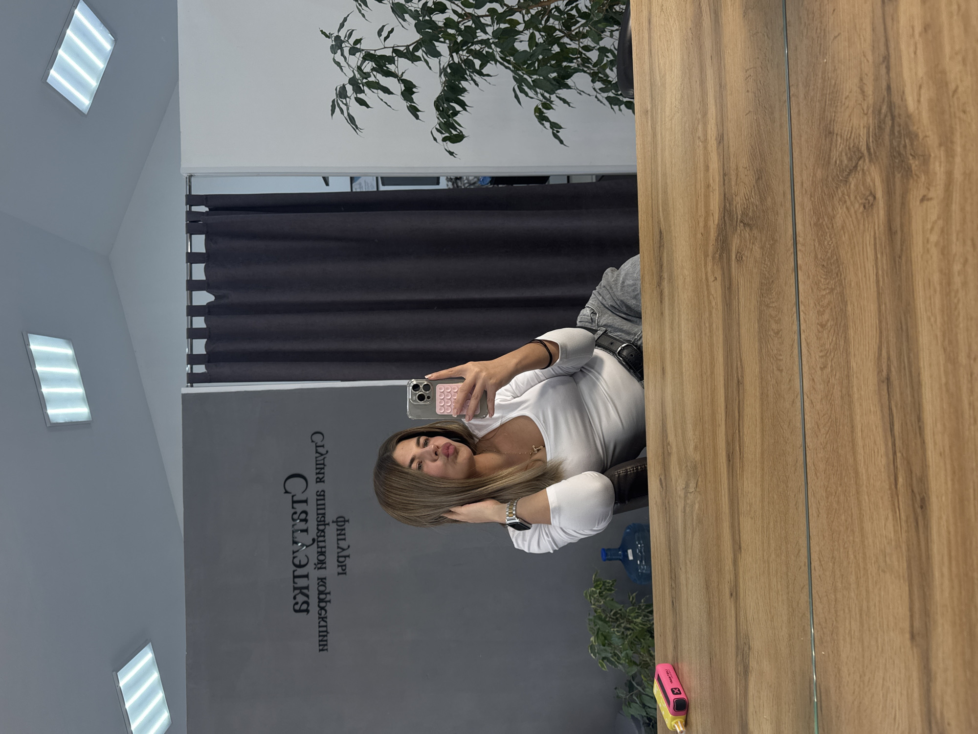 The image shows a woman taking a selfie in a mirror inside what appears to be a beauty salon. The reflection reveals a grey wall with Cyrillic text, possibly advertising the salon services, and a window with plants. The woman is holding a smartphone with a pink case, wearing a white top and watch. Her hair is styled with highlights. In the background, there's a large water bottle and a vaping device. The text on the wall indicates the location is likely a salon in Chaikovsky, Russia.
