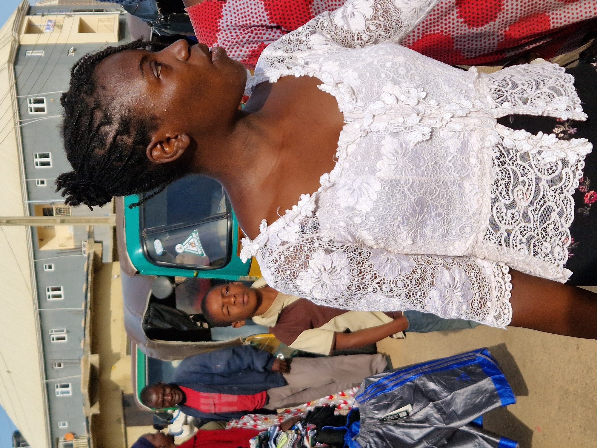 In Kabong, Nigeria, a group of people are gathered outdoors, likely at a marketplace. In the foreground, a woman with dark skin and braided hair is looking to the right. She is wearing a white lace blouse with intricate floral patterns and a black skirt. To her left, a young boy with dark skin is looking to the right, wearing a brown and tan collared shirt. Behind him, a man with dark skin and a red t-shirt under a dark blue jacket is looking forward. A three-wheeled auto-rickshaw is parked behind the man. In the background, a light-colored building with several windows is visible. The ground is paved.