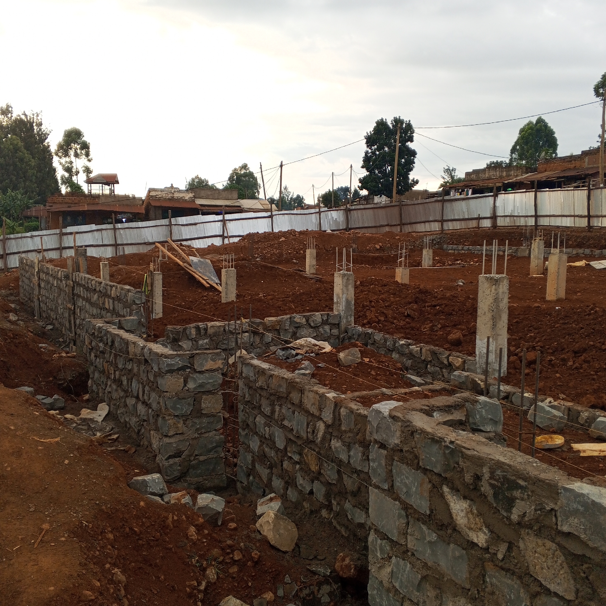 This image captures a construction site, likely in the early stages of building a new structure. The foreground features newly laid stone foundations, forming partial walls and outlines of rooms, built with irregularly shaped grey stones and mortar. String lines are visible along these walls, indicating precision in construction. Surrounding these foundations is abundant reddish-brown soil, a result of excavation.
Several concrete pillars, some topped with exposed rebar, are strategically placed throughout the site, suggesting future vertical support for the building. In the mid-ground, a corrugated metal fence acts as a boundary, behind which more excavated earth and additional concrete pillars can be seen. Wooden planks are also scattered on the disturbed ground.
The background reveals a mix of older buildings with corrugated iron roofs, indicative of a semi-urban or rural settlement. Lush green trees punctuate the skyline, and utility poles with power lines traverse the upper mid-ground. The sky is overcast and bright, suggesting either early morning, late afternoon, or simply a cloudy day.
The scene depicts ongoing development, with the visible elements pointing towards a multi-story construction project. No people are visible, but the active state of the site is evident. This setting is consistent with the provided location context of Borabu Chitago ward, Kenya, given the architecture and earthy landscape. There is no visible text in the image.