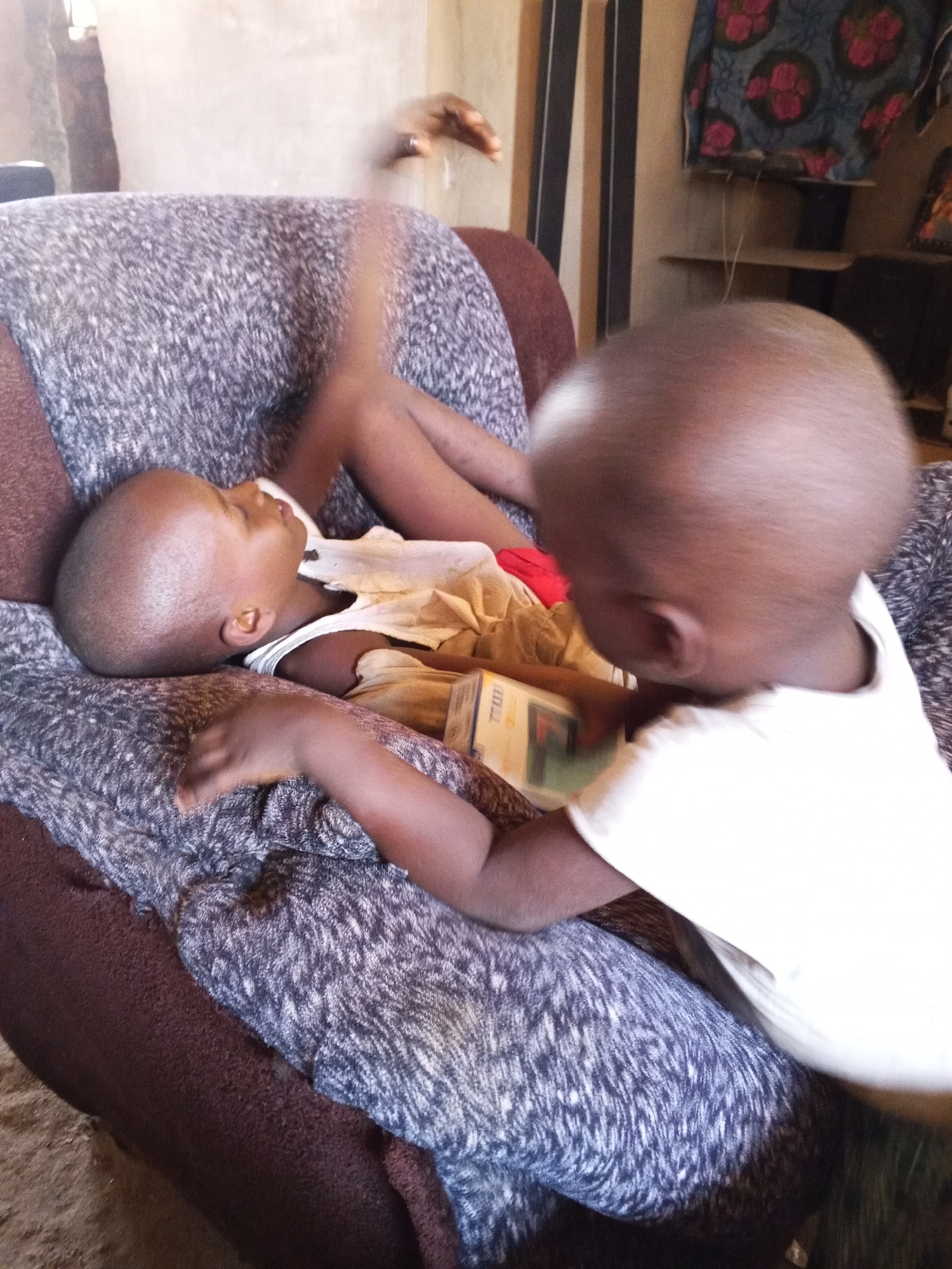 The image captures two young children indoors, likely in a home setting. One child, with dark skin and a shaved head, lies on their back on a textured sofa or armchair, wearing a light-colored tank top and possibly another garment. Their eyes appear closed or looking upwards, suggesting they are resting or asleep.
Leaning over this child is another young child, also with dark skin and a shaved head, wearing a white t-shirt. This second child's head is partially blurred, but they appear to be looking down at or interacting with a small rectangular object, possibly a book or a box, held near the first child's chest. The object has some indistinct text or branding, with faint letters that might be "TIDI" or "71DD" visible on its surface.
The sofa features a dark brown frame and a mottled grey and white fabric covering. In the background, bright light streams from what appears to be a window or doorway. Further back, a wall with vertical dark stripes is visible, along with a shelf or ledge. A piece of fabric, possibly a curtain or tapestry, with a repeating pattern of red floral designs against a dark background, hangs on the wall. A blurry hand, possibly an adult's, is visible in the upper background, suggesting another person might be nearby. The scene appears to take place during the daytime. The overall atmosphere seems calm and domestic.