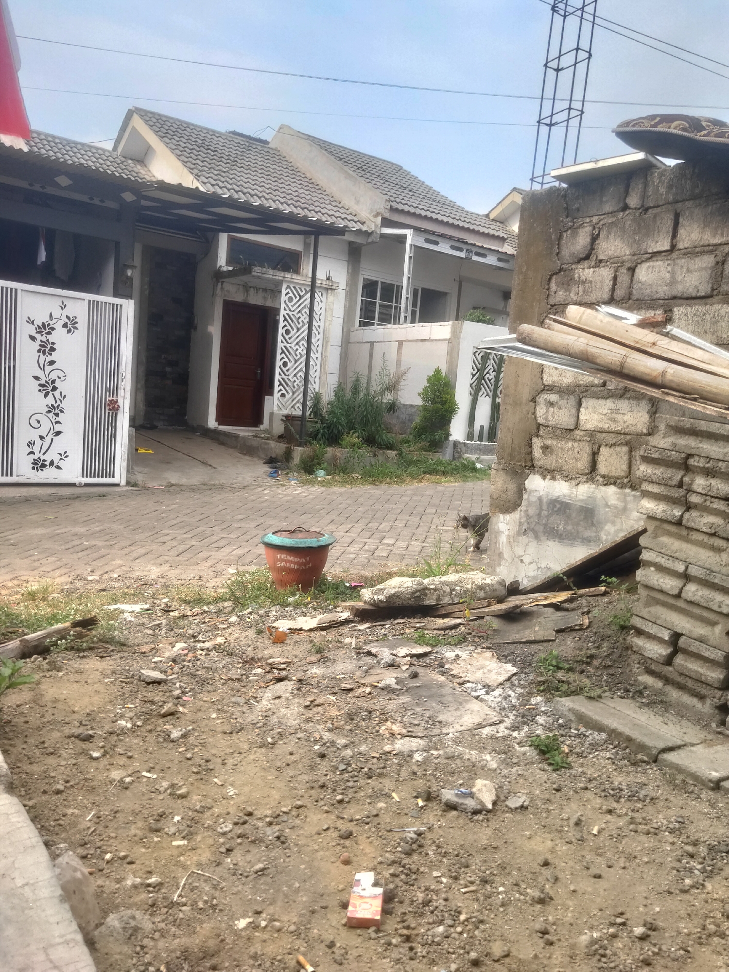 The image shows a residential area in Antirogo, Indonesia, with a focus on a partially constructed or renovated section of a property. In the foreground, the ground is covered with dirt, small rocks, and scattered debris, including cigarette butts and what appears to be a pack of cigarettes. A planter with the text "TEMPAT SAMPAH" (meaning "trash bin") is visible in the middle ground, near a paved walkway. Behind the planter, a cat is walking on the paved area. Further back, a white metal gate with a floral design is open, leading to a house with a dark brown door and tiled roof. The house appears to be a single-story structure with modern architectural elements, including decorative grilles on the walls. To the right of the house, a wall constructed from rough concrete blocks is prominent, with stacked bamboo poles and other building materials leaning against it. The overall scene suggests a residential environment undergoing some form of construction or maintenance, with elements of everyday life, such as the planter and the cat, integrated into the scene. The sky above is clear and light blue.
