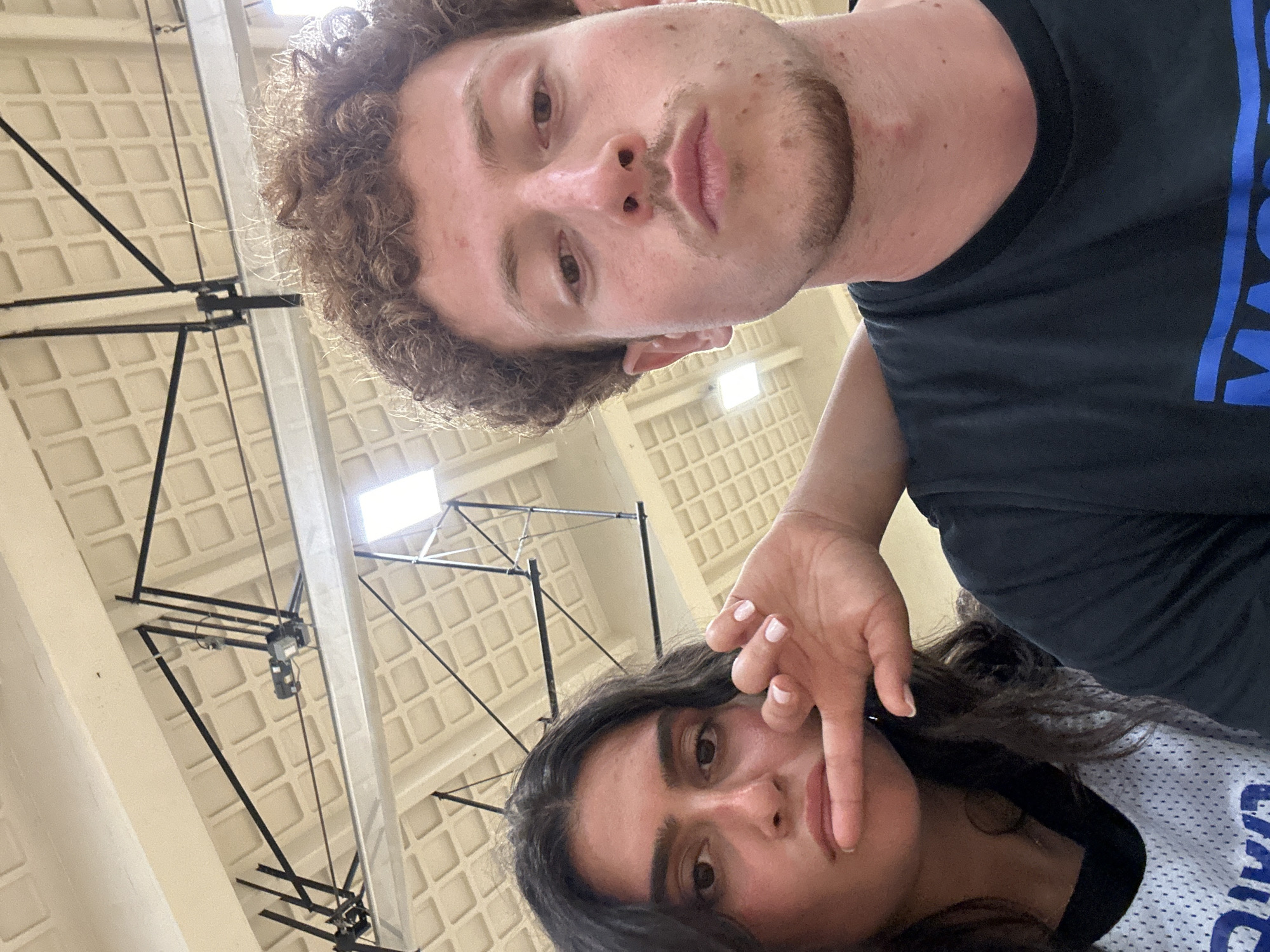 The image shows two individuals in what appears to be an indoor sports facility, possibly a gymnasium, located in College Park, United States of America. A male with curly brown hair and a light beard is positioned closer to the camera, wearing a black t-shirt with blue lettering. Below him, a female with long, dark hair is visible, wearing a white and blue basketball jersey. Her hand is raised near her face. The background shows a high ceiling with a grid-like pattern and overhead lighting, along with black metal structures that seem to be part of the gymnasium's equipment.
