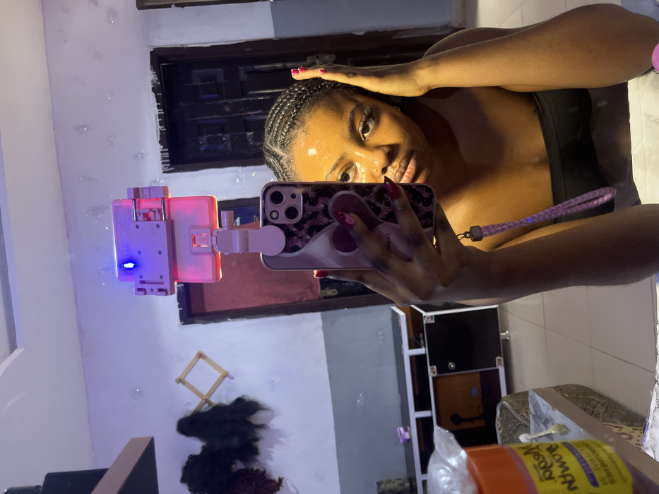 A young adult female with dark skin and dark braided hair (cornrows) is depicted in a mirror selfie, taken indoors in Gbongan, Nigeria. She wears a dark, strapless top. Her right hand, with red manicured nails, holds a smartphone in a pink and black leopard-print case, which is attached to a device emitting pink and blue light, likely a selfie stick with an integrated ring light. A pink wrist strap hangs from the phone. Her left hand is positioned near her left ear. She maintains a neutral expression, looking directly towards the camera. The reflected background reveals a domestic interior with light-colored walls, a dark paneled door, and a light-colored tiled floor. On the left, dark hair extensions or wigs hang from a wooden rack on the wall, above a dark cabinet with lighter panels. An orange container, possibly a cosmetic product, with partially visible text "Nodra," is reflected in the lower left foreground.