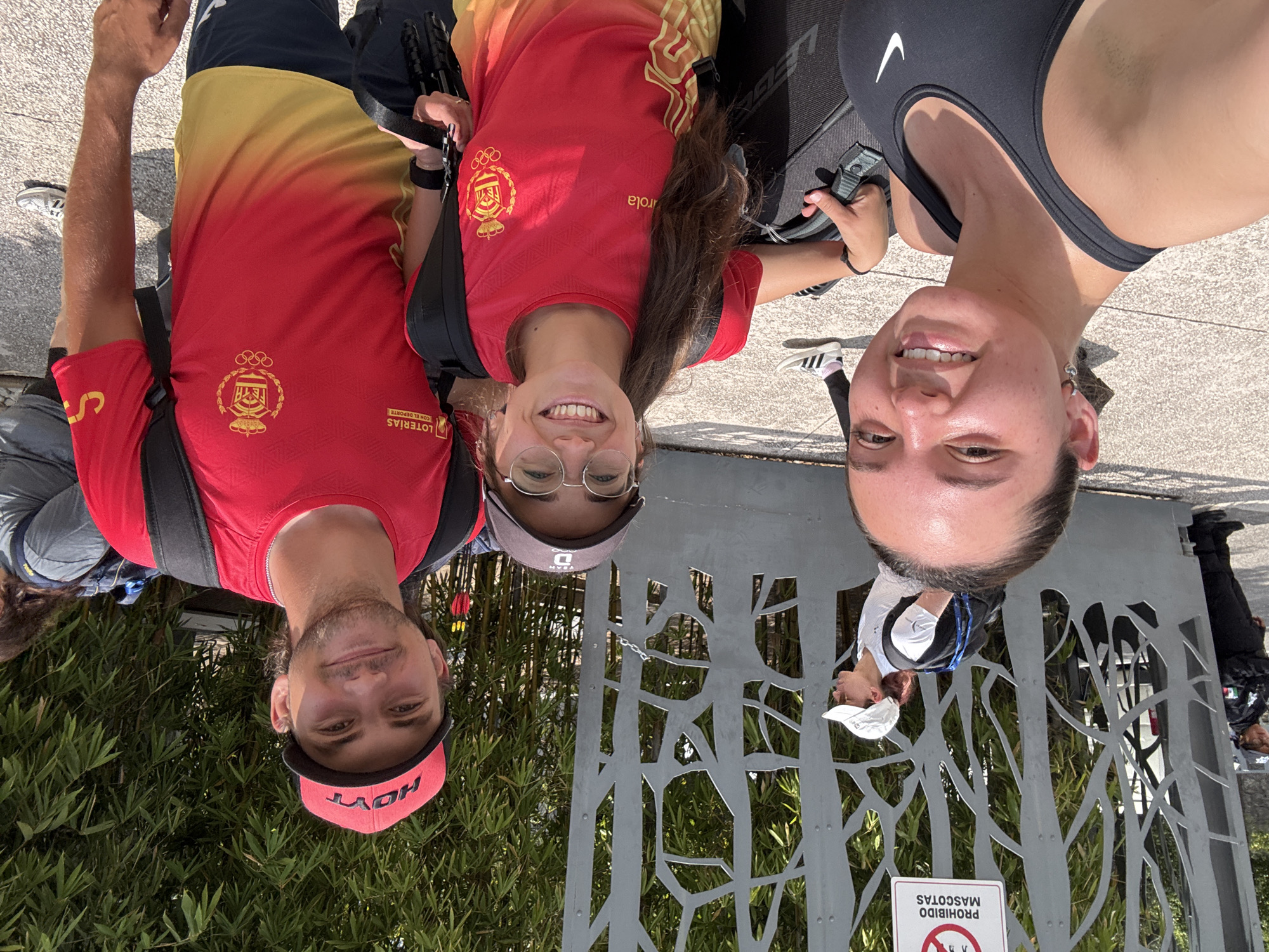 Three individuals are posing for a selfie in front of a gate with a tree-like design. The person on the left wears a red and yellow t-shirt, a red cap with "HOYT" on it, and has a backpack. The person in the middle also wears a red t-shirt with an Olympic logo, glasses, and a cap. The person on the right wears a black sports top. The gate has a sign that reads "PROHIBIDO MASCOTAS". Behind the gate, bamboo plants can be seen. The selfie appears to be taken in the city of Puebla, Mexico.