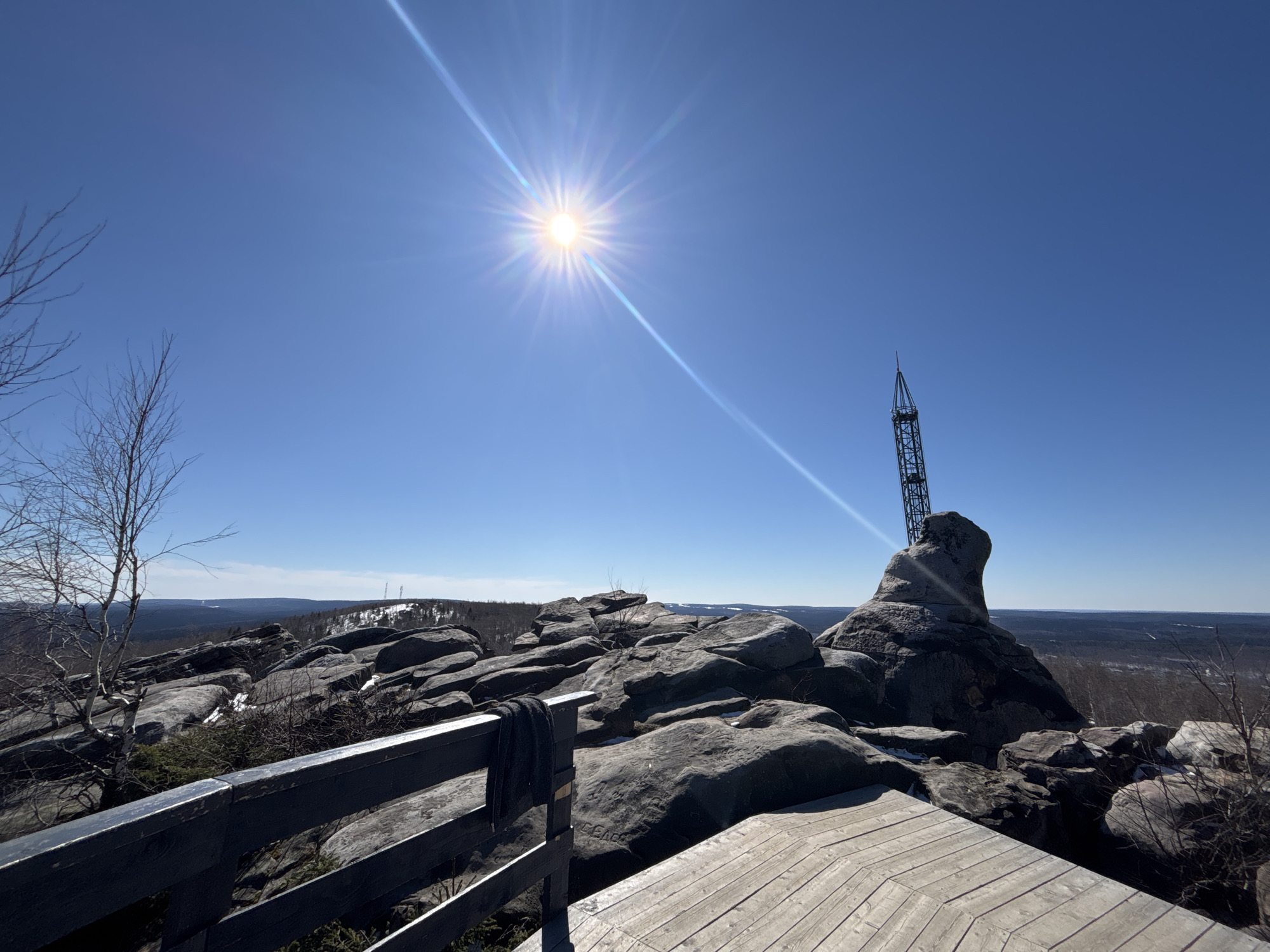 The image shows a sunny, daytime scene from a high vantage point in Gubakha, Russia. Dominating the foreground are large, grey rock formations. A wooden deck with a railing is visible, suggesting a constructed viewing platform. A dark grey garment hangs over the railing. A tall metal structure stands atop one of the rocks, possibly a communications tower or observation point. Beyond the rocks, a landscape of forested hills stretches into the distance, with patches of snow still visible. The sky is clear and bright blue, with the sun shining intensely. Bare branches of trees are visible in the lower left.