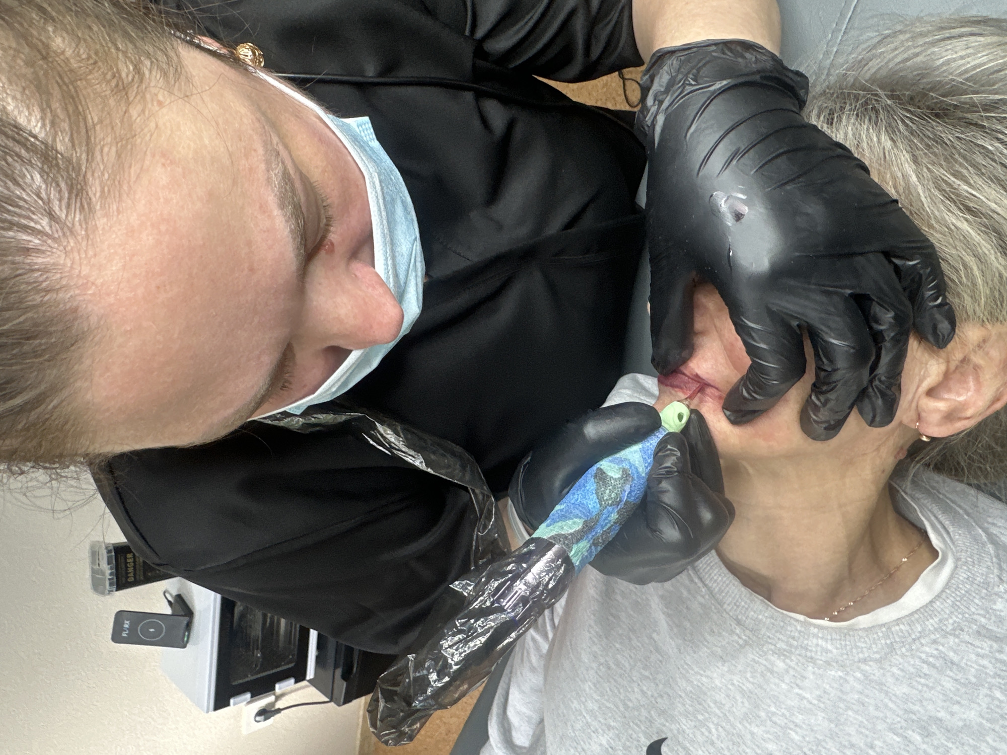 A tattoo artist is applying permanent makeup to a female client's lips. The artist wears a black uniform and a blue surgical mask. Black gloves cover the artist's hands, which hold a tattoo pen wrapped in a protective plastic sleeve and a camouflage bandage. The client wears a gray sweatshirt and a gold necklace. The location appears to be an indoor studio with white walls. It is not possible to identify the location as Chaikovsky, Russia, from the image.