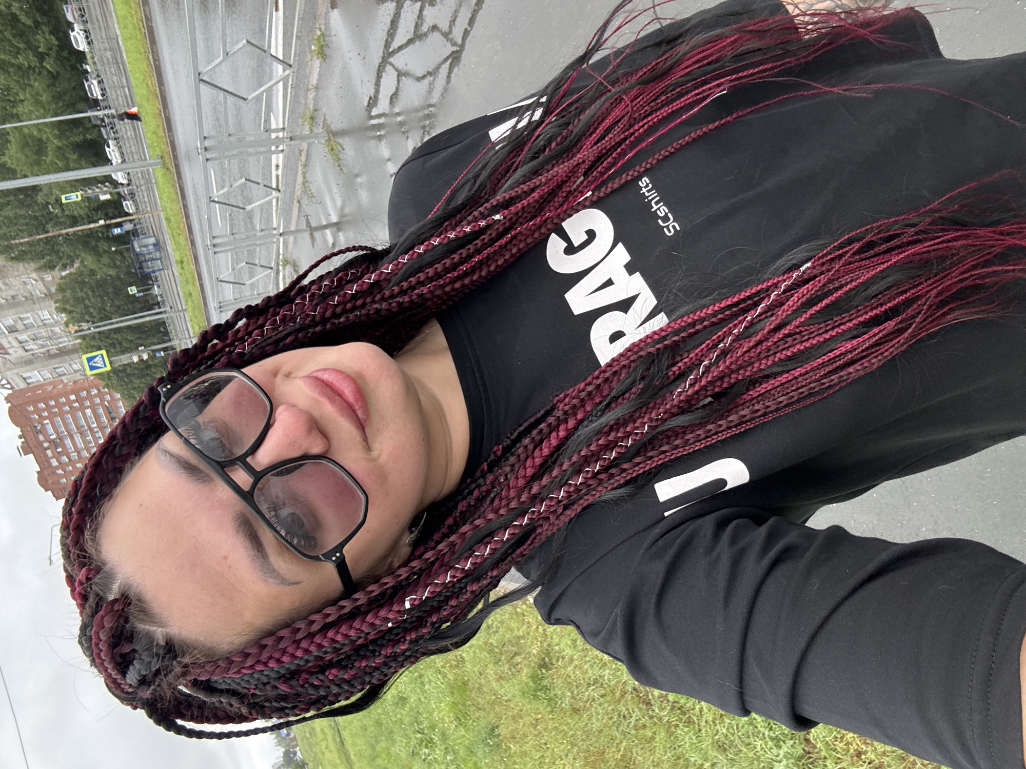 The image shows a woman with dark red and black braided hair, wearing large black-framed glasses and a black t-shirt with white lettering that appears to say "RAG SCshirts". She is taking a selfie outdoors in what appears to be a city environment. The background includes a wet asphalt road, a metal fence, green grass, trees, and buildings. Traffic signals and parked cars are visible in the distance, suggesting a street or roadside location. The overall atmosphere is overcast. While the specific location in Tolyatti, Russia, cannot be determined solely from the visual information, the presence of Cyrillic lettering on street signs or buildings would be a strong indicator.