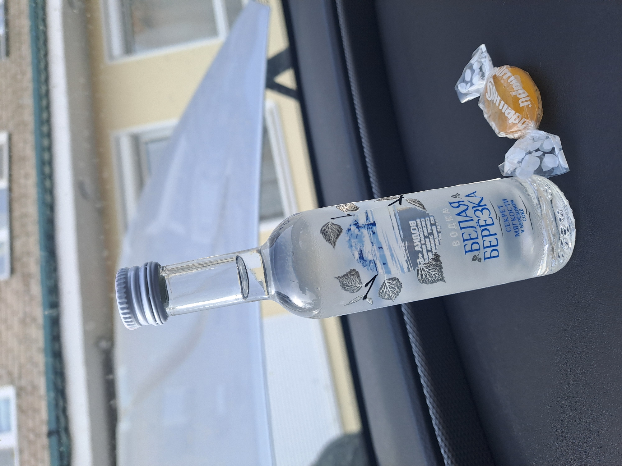 The image displays a clear glass miniature bottle of "Белая Березка" (Belaya Berezka) vodka, distinguished by a label featuring silver birch leaves and a blue/white landscape motif. The bottle, topped with a silver screw cap, exhibits condensation on its exterior, indicating cold temperature. Adjacent to the bottle are two individually wrapped candies; one is yellow with visible "Sim-Sim" branding, while the other is partially obscured but also appears wrapped in clear plastic. These items rest on a dark, textured surface, consistent with a vehicle's dashboard or console.
Through the foreground windshield, the background reveals a light-colored building in the city of Elektrostal, Russia. The building features multiple white-framed windows, with an upper section constructed of light brown brick, beneath which is a green trim or gutter. The lower visible section of the building presents a lighter yellow or beige wall surface. A large, light-colored, possibly white, sheet or tarp is partially visible in front of the building, creating a bright reflective area in the background. No individuals or explicit actions are present in the frame.