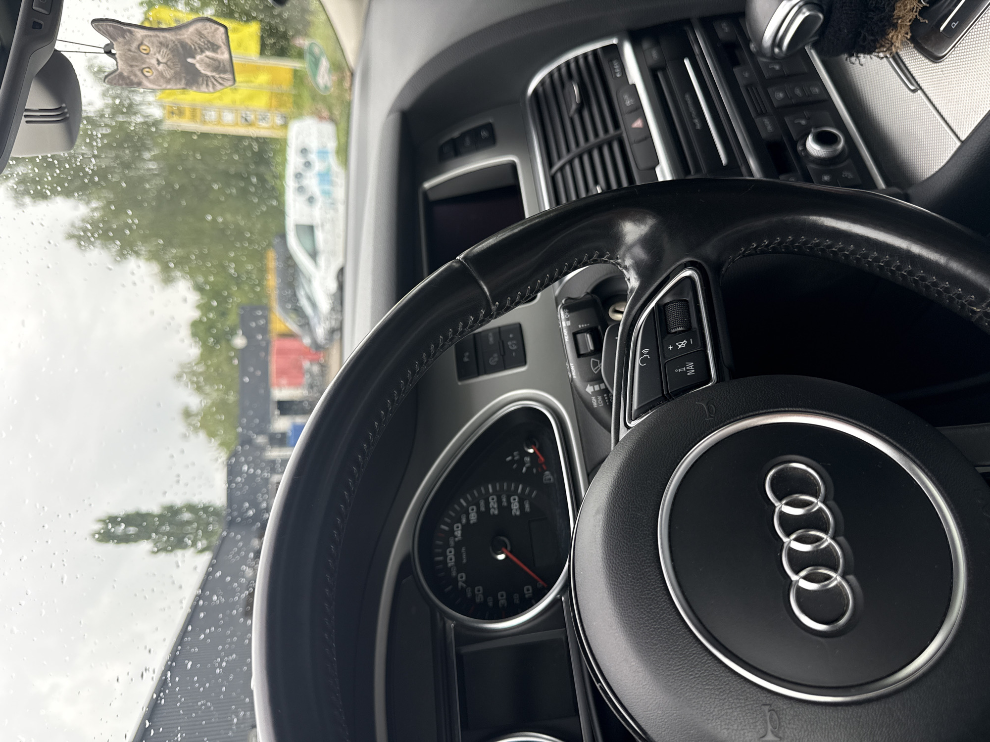 The image shows the interior of an Audi car, viewed from the driver's seat. The steering wheel, with the Audi logo, is prominently displayed. The dashboard features a speedometer and other instruments. A car air freshener shaped like a cat is hanging from the rearview mirror. The windshield is covered in raindrops, suggesting it is raining outside. In the background, through the windshield, a building and trees are visible, along with parked vehicles, including a white van. The location is the city of Vellmar, Germany.
