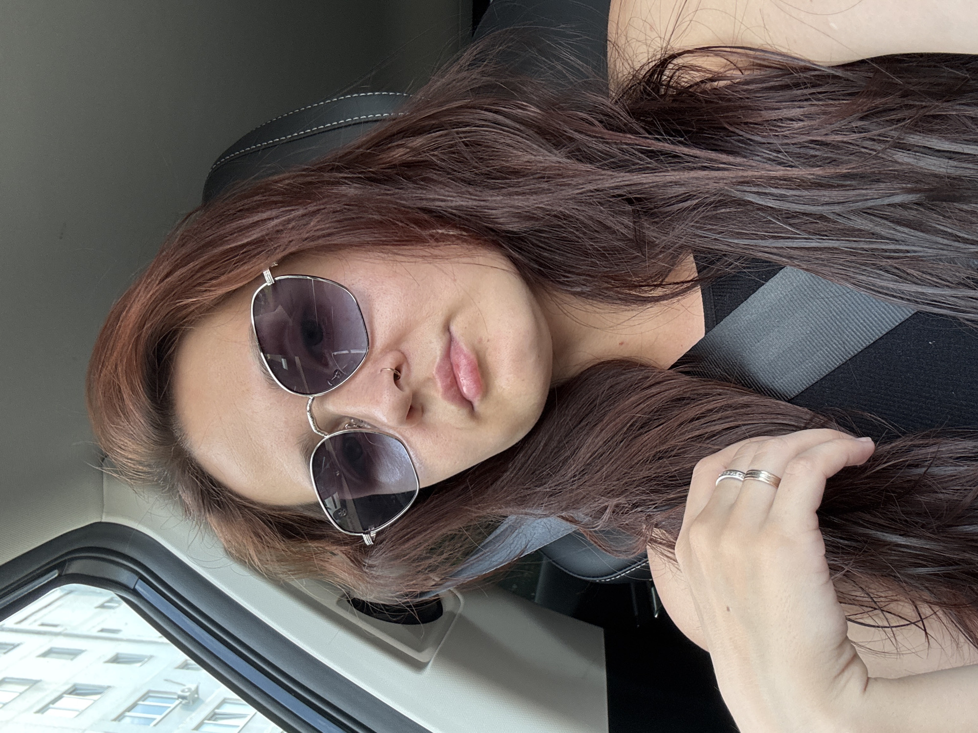 A woman is captured from the chest up, inside a car. She is wearing sunglasses and a black tank top. Her hand, adorned with two rings, rests on her hair. The car's interior, including a seatbelt and part of the car door, is visible. Outside the car window, a multi-story building, typical of Naberezhnye Chelny, Russia, can be seen.