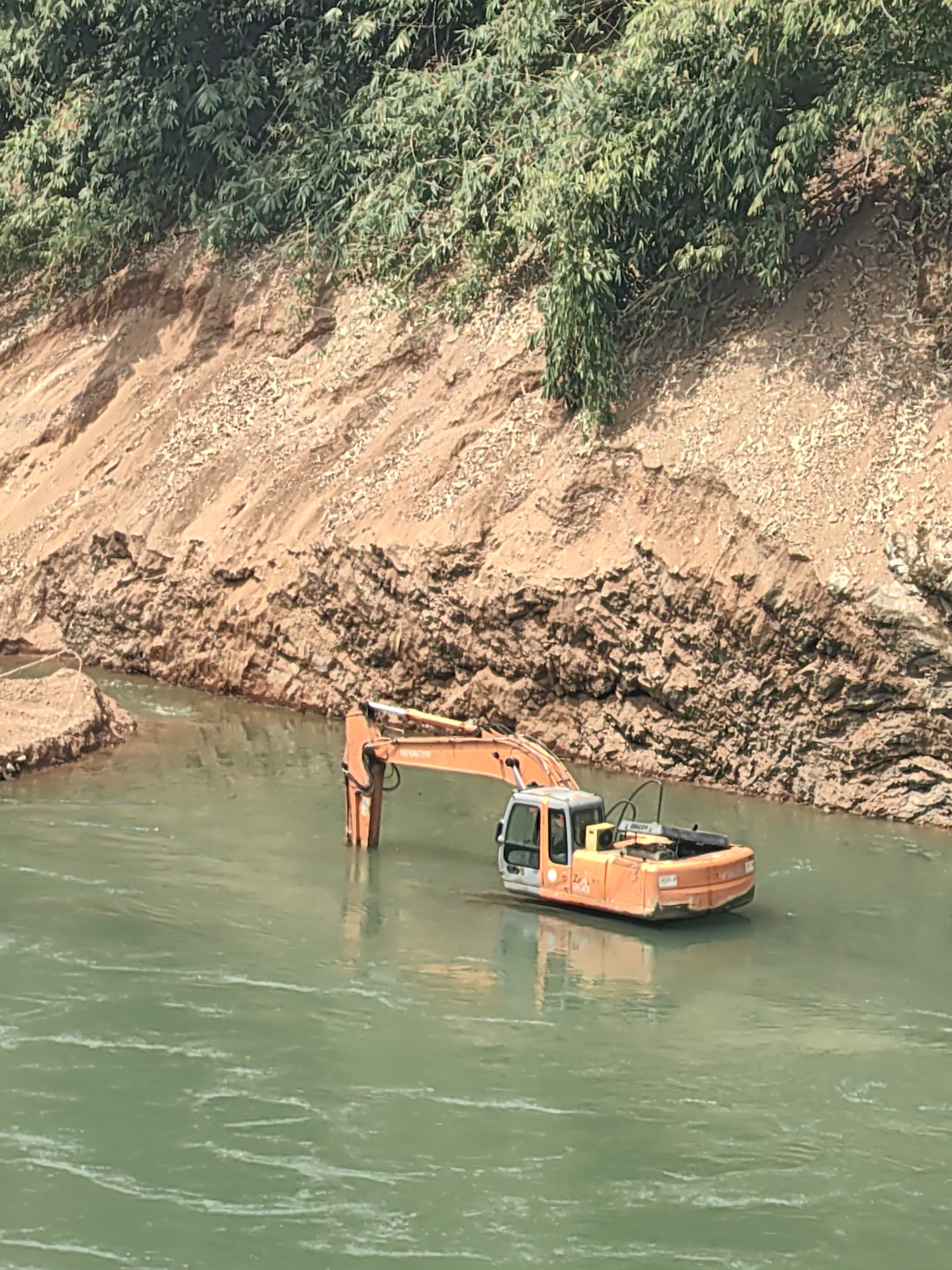 An orange Hitachi excavator sits mostly submerged in a green river in Bảo Lạc, Vietnam. The excavator’s boom is extended upwards. A brown, dirt cliff with vegetation along the top rises on the right side of the river.