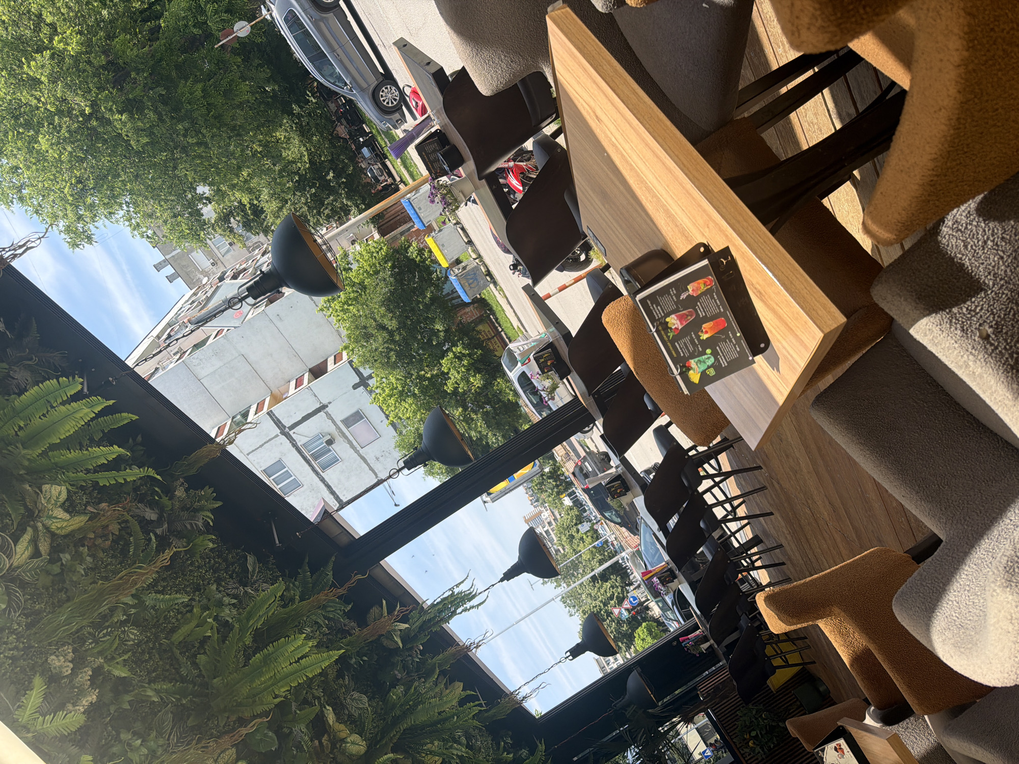 This image depicts the outdoor or semi-outdoor seating area of a cafe in an urban setting, specifically in Shumen, Bulgaria, during clear, sunny daytime conditions. The foreground and midground feature multiple cafe tables with light wooden tops and dark metal bases, surrounded by various seating options including dark chairs and upholstered armchairs in grey and brown textured fabric. A black stand with a colorful beverage menu, including "Лимонади" (Lemonades) in Bulgarian, is visible on one of the tables. The overhead structure of the cafe is densely covered with a canopy of lush green foliage, composed of ferns and other plant types. Several black industrial-style pendant lamps hang from this green ceiling. Beyond the cafe, a city street is visible with cars, including a grey SUV, a white van, and a red motorcycle, parked and in motion. The sidewalk features blue and yellow public waste bins. The street is lined with large, green trees, which partially obscure multi-story residential buildings characterized by light-colored facades and visible air conditioning units. Street signs are also present. No individuals are explicitly visible within the scene.