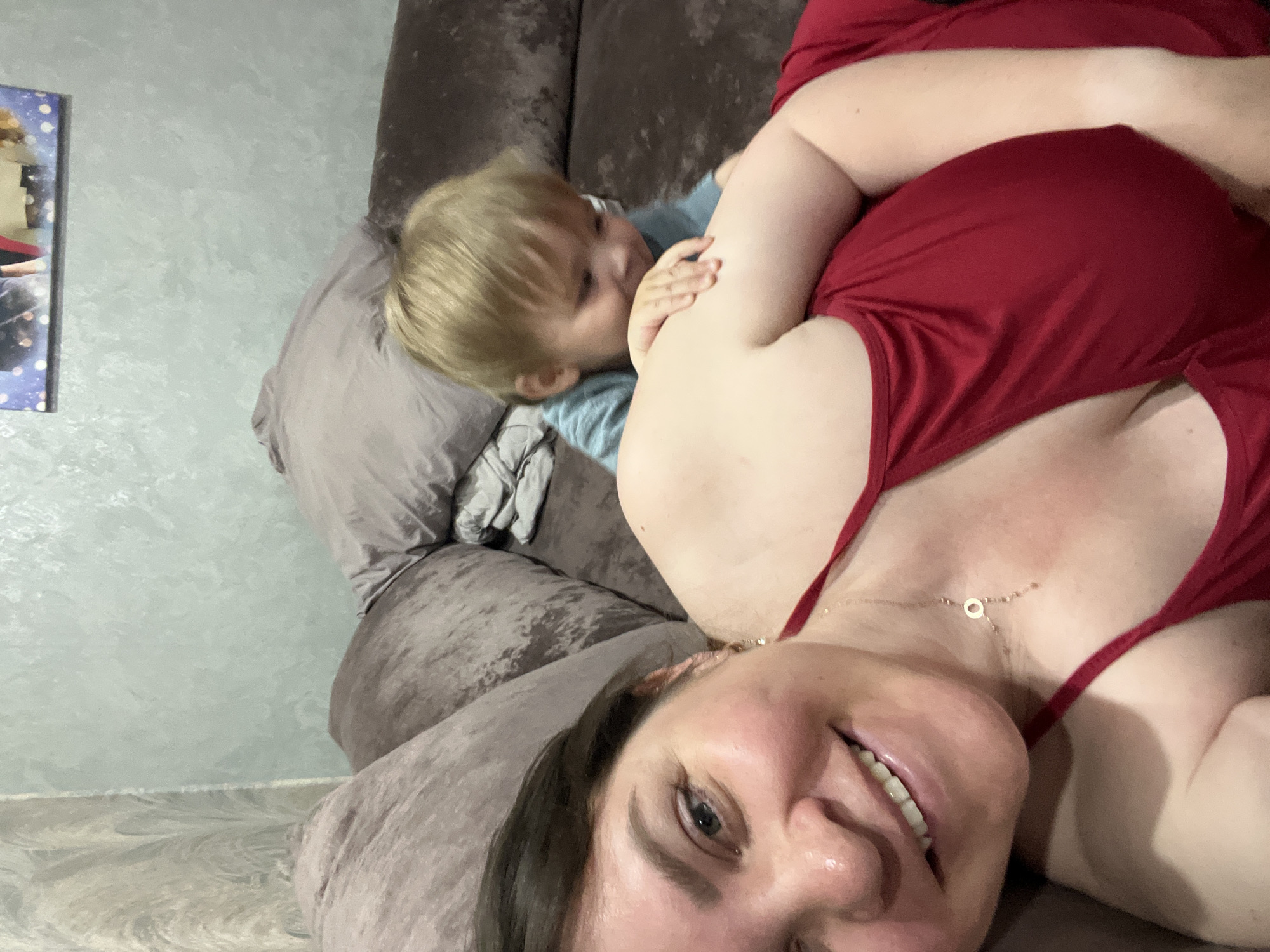 A woman and a child are on a couch. The woman is in the foreground, smiling at the camera. She has brown hair and is wearing a red dress. The child, a toddler with blond hair, is behind her, looking down. The background shows a textured wall and a framed picture. The scene appears to be an indoor setting, likely a living room. The location is specified as Noviy, Russia.