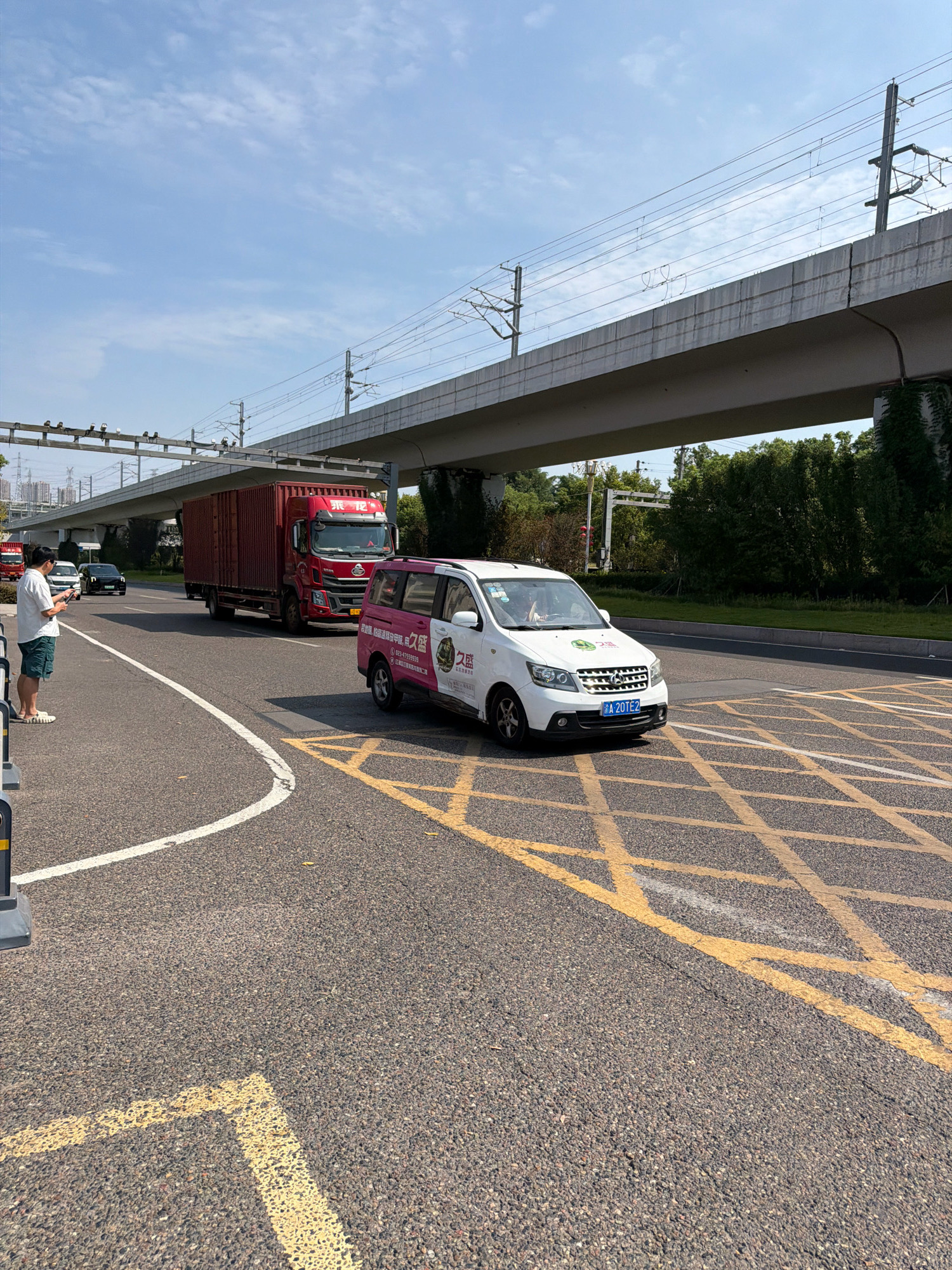 In Jiangjin, China, a person stands on the side of a road, looking at their phone. A white van with pink and green decals, and a blue license plate reading "A 20TE2", is parked on a section of road marked with yellow criss-cross lines, indicating no stopping or waiting. To the left of the van, a red semi-truck is moving forward. In the background, a large elevated structure, possibly a highway or a train track, runs across the scene, supported by concrete pillars. The sky is clear blue with some clouds. Trees and buildings are visible in the distance.