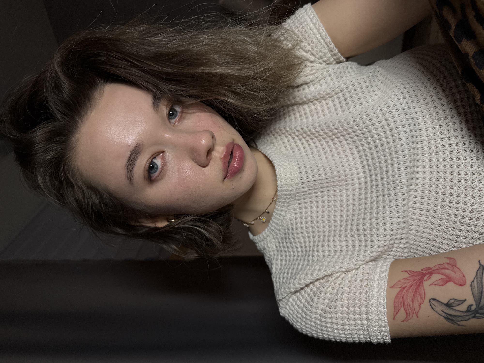 A close-up, sideways-oriented portrait captures a young woman reclining in an indoor setting. She has light skin, medium-length wavy light brown hair, and prominent blue-green eyes that gaze directly at the viewer. Her lips are full and have a soft pink hue. She is wearing a light-colored, possibly cream or off-white, waffle-knit long-sleeved top. A delicate gold chain necklace adorned with a small charm, resembling a daisy, is visible around her neck.
On her right forearm, two distinct fish tattoos are apparent: one intricately outlined in red, and the other in black and grey. A dark, indistinct background suggests low, soft indoor lighting, which highlights areas of her face like her forehead and nose. A section of brown and black animal print fabric is partially visible on the far right of the frame, hinting at another piece of clothing or decor. The woman maintains a calm and direct expression while posing for the photograph. No visible text is present in the image.