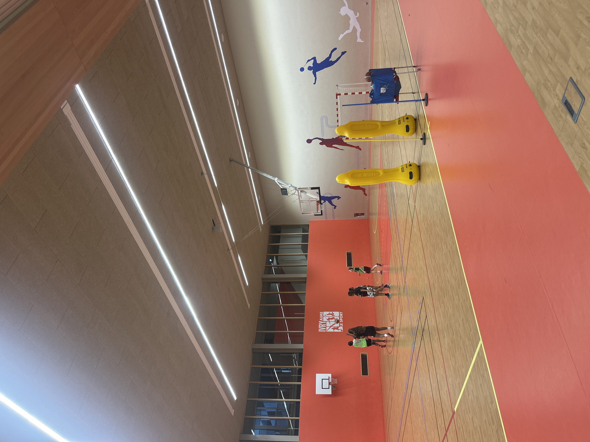 A brightly lit indoor gymnasium in Ivry-sur-Seine, France is photographed from an elevated perspective. Several people are present on the wooden floor, which is marked with various colored lines and sections of solid red. The people appear to be engaged in sports activities. Various pieces of sports equipment, including yellow cylindrical objects, a net with blue equipment behind it, and basketball hoops, are located throughout the space. Artwork depicting athletes is affixed to the upper walls. Large windows are visible, as well as rows of overhead lights. A logo of "Ivry aime le Sport" is visible on the wall.