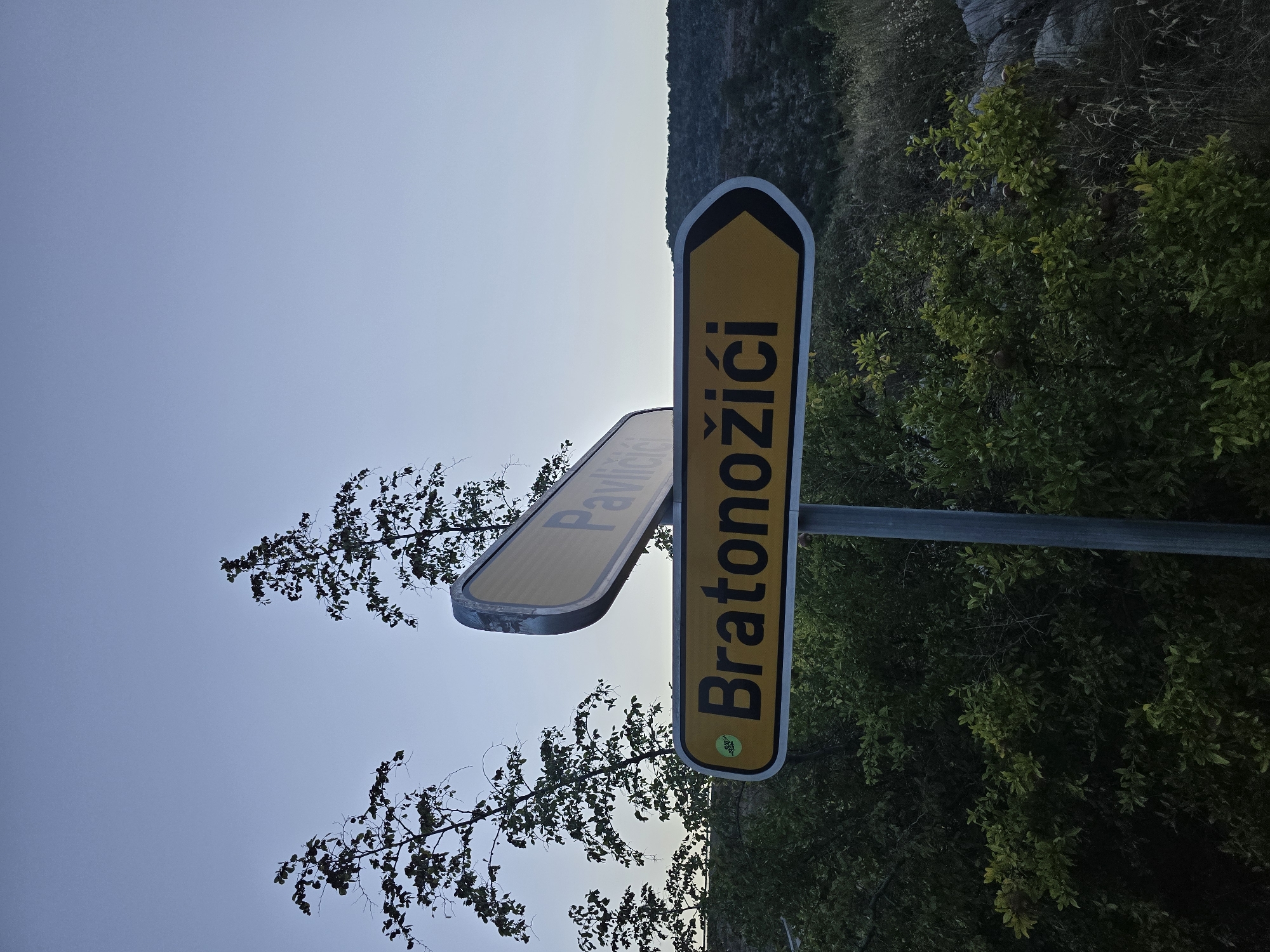 A street sign in Klopot, Montenegro, displays directions to "Bratonožići" and "Pavlici". The "Bratonožići" sign is yellow with black text and an arrow pointing to the right. Above it, a second yellow sign, partially obscured, reads "Pavlici". Both signs are mounted on a metal pole. The scene is set against a backdrop of trees and a rocky, hilly landscape under a clear, pale blue sky.