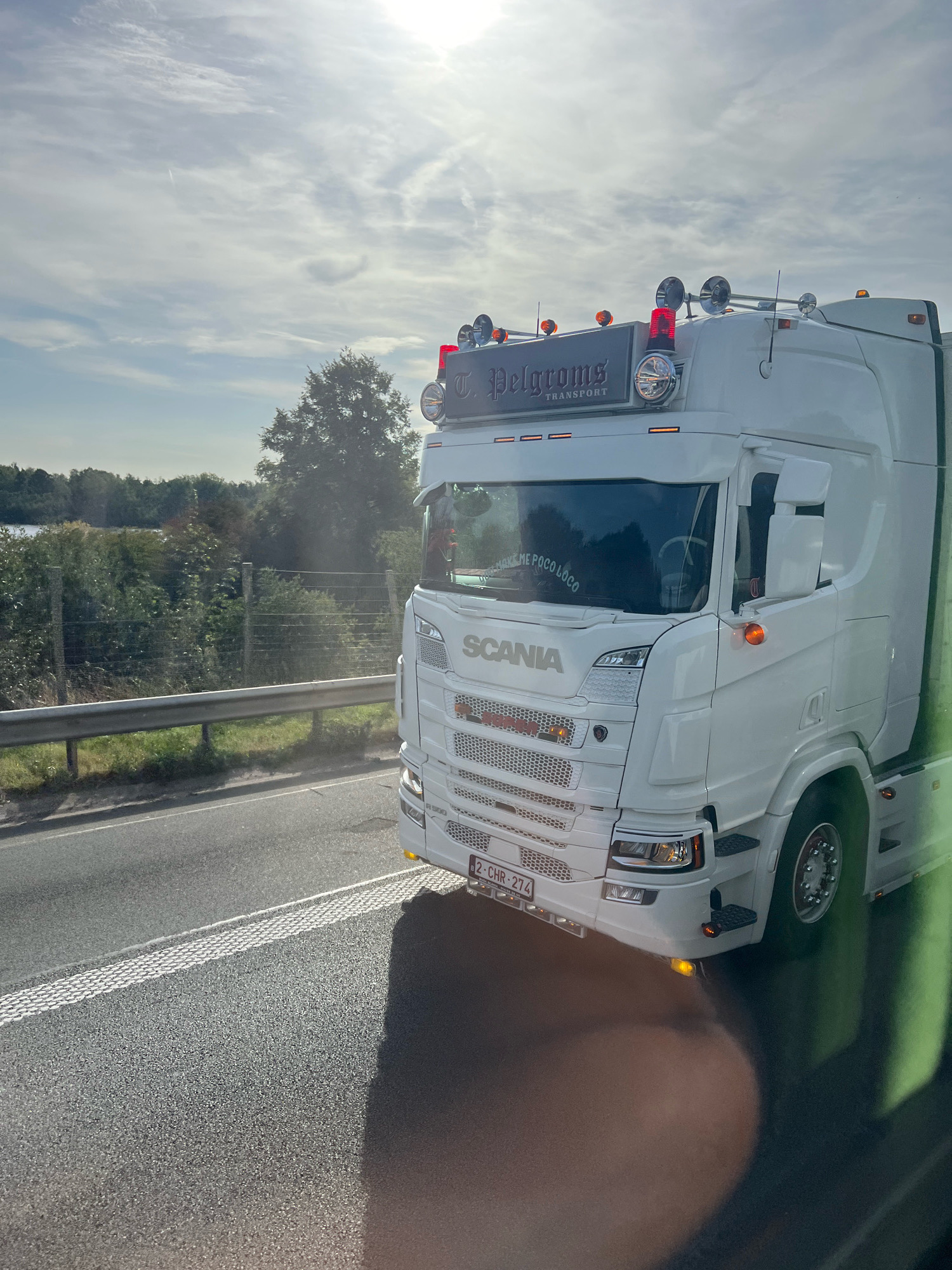A white Scania truck with the name "T. Pelgroms Transport" is driving on a road. The truck has red and orange lights on its roof. There is a fence and trees on the left side of the road, and a body of water in the distance. The sky is cloudy and the sun is shining through. The license plate of the truck is visible and reads "2-CHR-274". There is text on the windshield of the truck that reads "DON'T MAKE ME POCO LOCO". The scene appears to be a roadside in or around Mechelen, Belgium, given the license plate format.