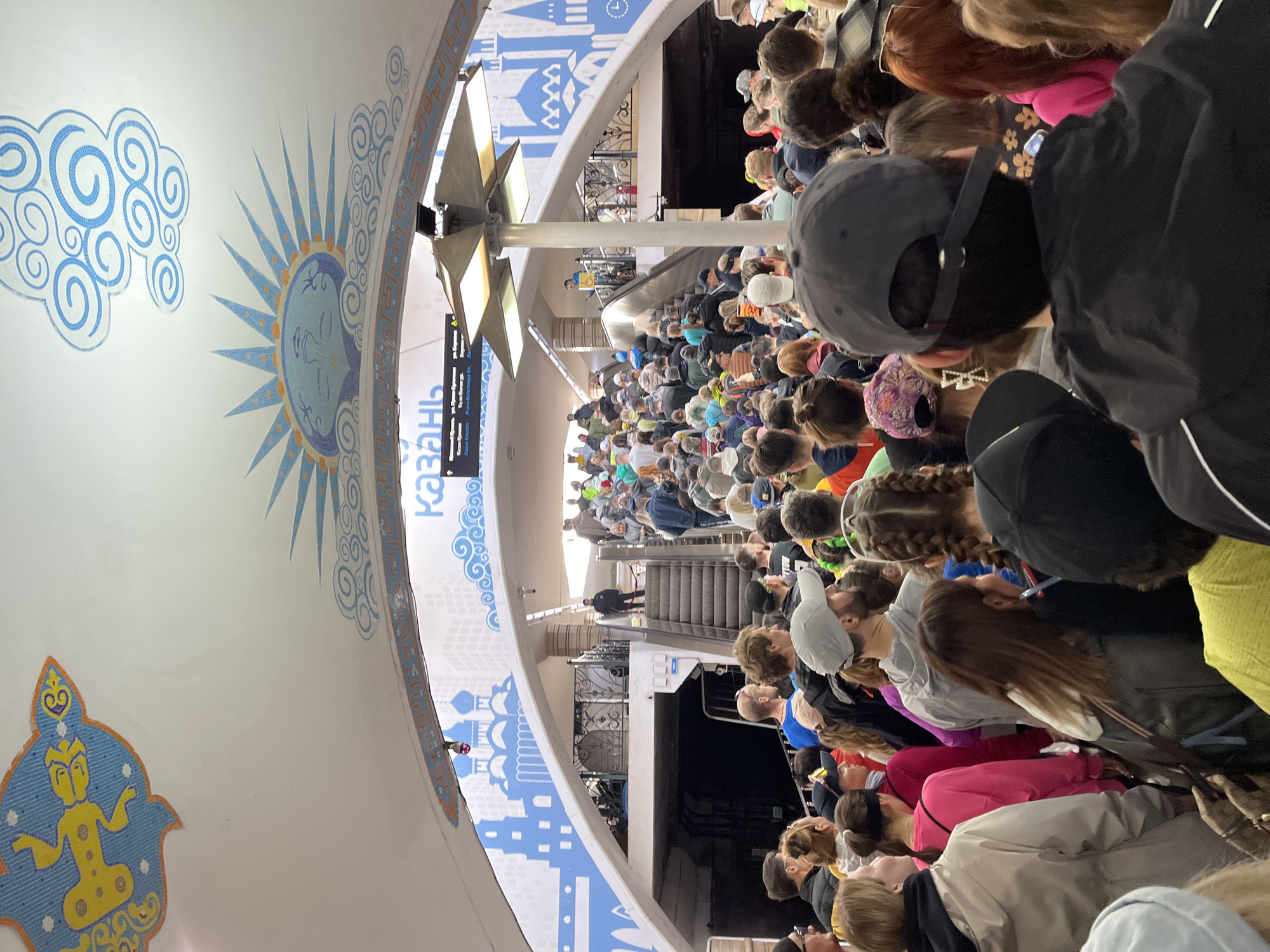 The image shows a crowded area inside the Kazan Metro in Kazan, Russia. A large group of people are gathered, appearing to wait in a line, likely at a station entrance or near escalators. The interior architecture features ornate, white vaulted ceilings decorated with blue mosaic patterns, including representations of a sun, clouds, and stylized figures. The signage indicates the station name is "Kazan". The lighting is bright and artificial. People are dressed in various casual clothing. Some individuals are visible on escalators.