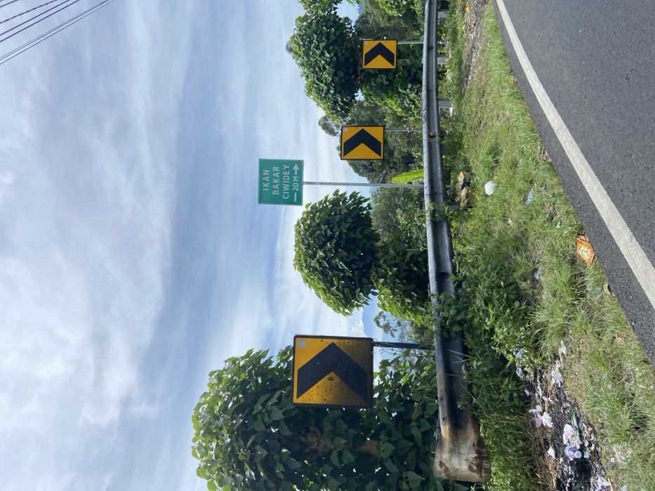 The image shows a winding road with a guardrail and a green sign that reads "IKAN BAKAR CIWIDEY -20M". The sign is located on the right side of the road. The road is paved and curves to the left. There are several yellow warning signs with black arrows indicating a sharp bend in the road. The surrounding vegetation is lush and green with trees lining the roadside. The sky is blue with white clouds. This location is likely a road in the hilly areas of Ciwidey, Rancabali, Indonesia.