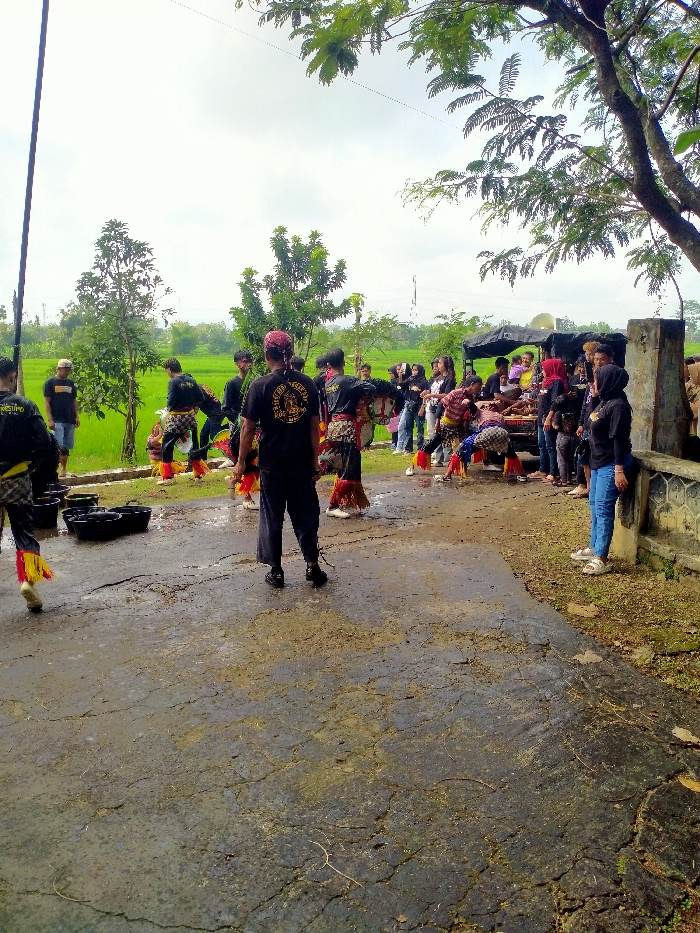 The image shows a group of people gathered in an open area in Kecamatan Kedawung, Indonesia. The group is comprised of 20 individuals, standing in a semi-circle around a truck, with some seated and others standing. Some people are wearing traditional clothing with yellow and red accents, while others are dressed in casual attire. Several people are interacting with each other while others are focused on the action happening in front of them. The scene is set in a rural environment with a grassy field and a dirt path leading to the group. There are several black plastic buckets scattered on the ground in front of the group. The trees are bare and the sky is overcast with a few wispy clouds. The overall feel of the image is calm and peaceful. The scene is a celebration of some kind, perhaps a cultural event or a religious ceremony. The image is a snapshot of a moment in time, and it captures the essence of community and tradition.