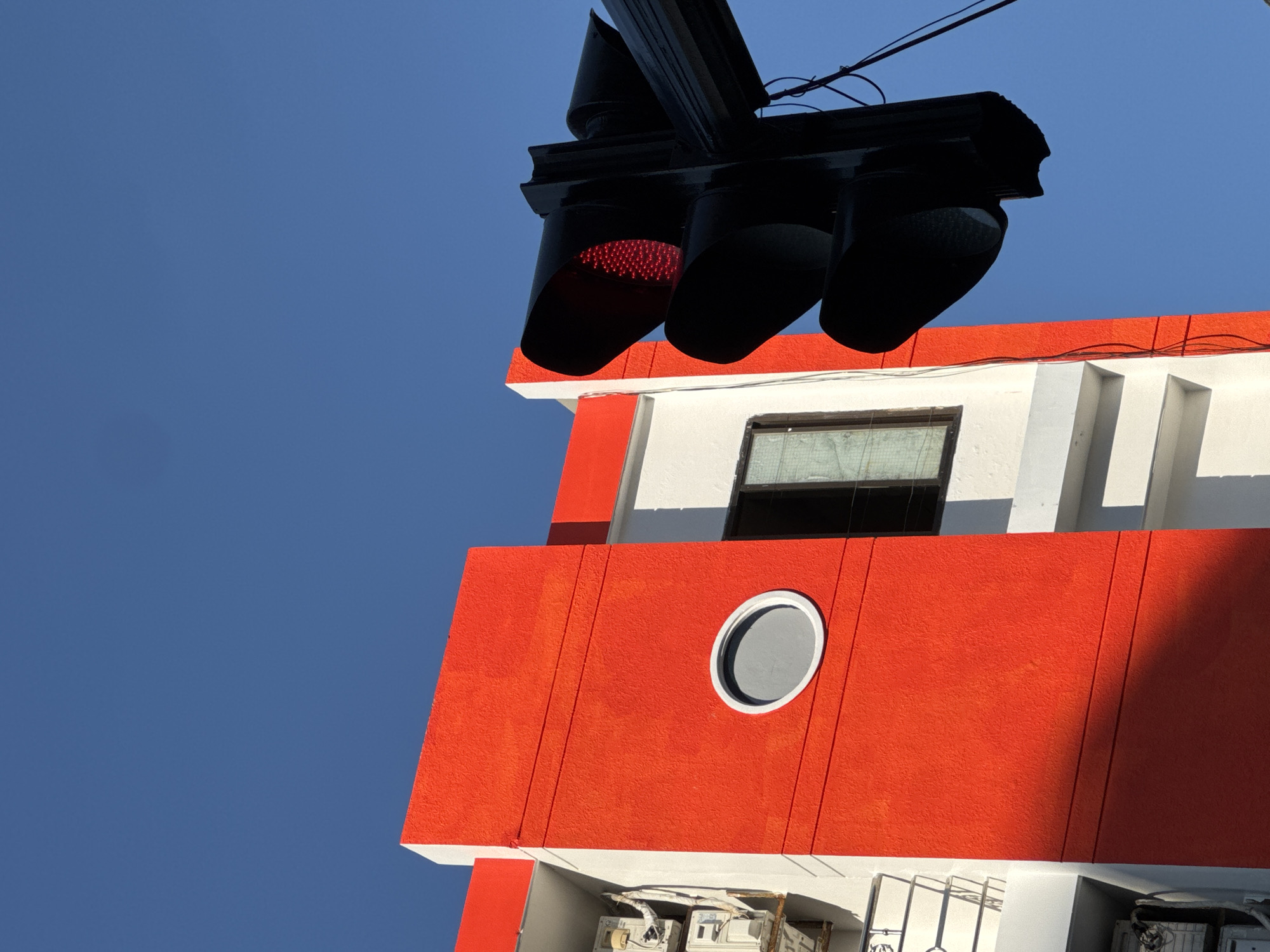 A traffic light with a lit red signal is positioned in the foreground against a bright blue sky. To the left of the traffic light, a section of a building with a striking orange facade is visible. The building has white trim and features a circular, grey-tinted window. The lower part of the orange facade is partially obscured by a shadow. The scene suggests an outdoor urban setting.
