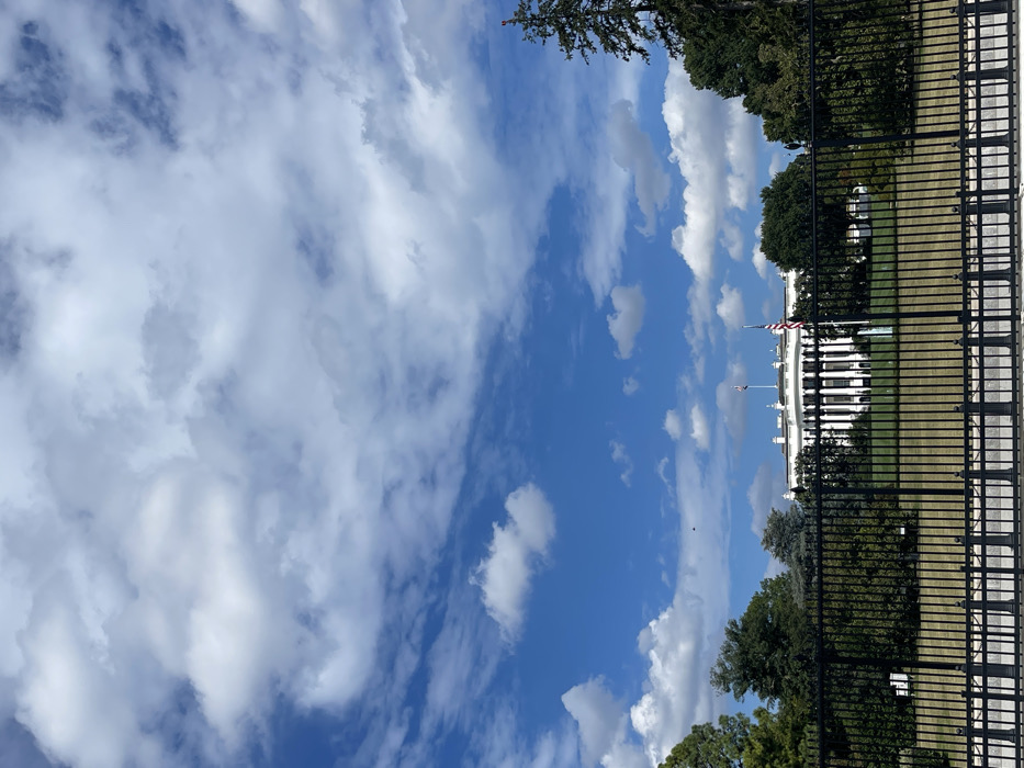 The image depicts the North Lawn view of the White House in Washington, D.C., United States, observed from outside its perimeter. In the foreground, a prominent black metal fence with vertical pickets and horizontal rails stretches across the width of the frame, resting on a concrete or stone base. Behind the fence, a manicured green lawn is visible, leading to the White House itself.
The White House, a white neoclassical building with a central portico featuring columns, dark windows, and a dark roof, occupies the midground. Two flagpoles are present: one taller pole on the lawn to the right of the building, flying an American flag, and another smaller flagpole on the building's roof, also flying a flag. Green deciduous and coniferous trees and shrubs line the perimeter behind the fence and flank the White House structure on both sides. A tall evergreen tree stands prominently on the far right. The upper two-thirds of the image consist of a clear blue sky sparsely populated with scattered white cumulus clouds. No individuals or discernible actions are present in the scene. The lighting indicates daytime conditions.