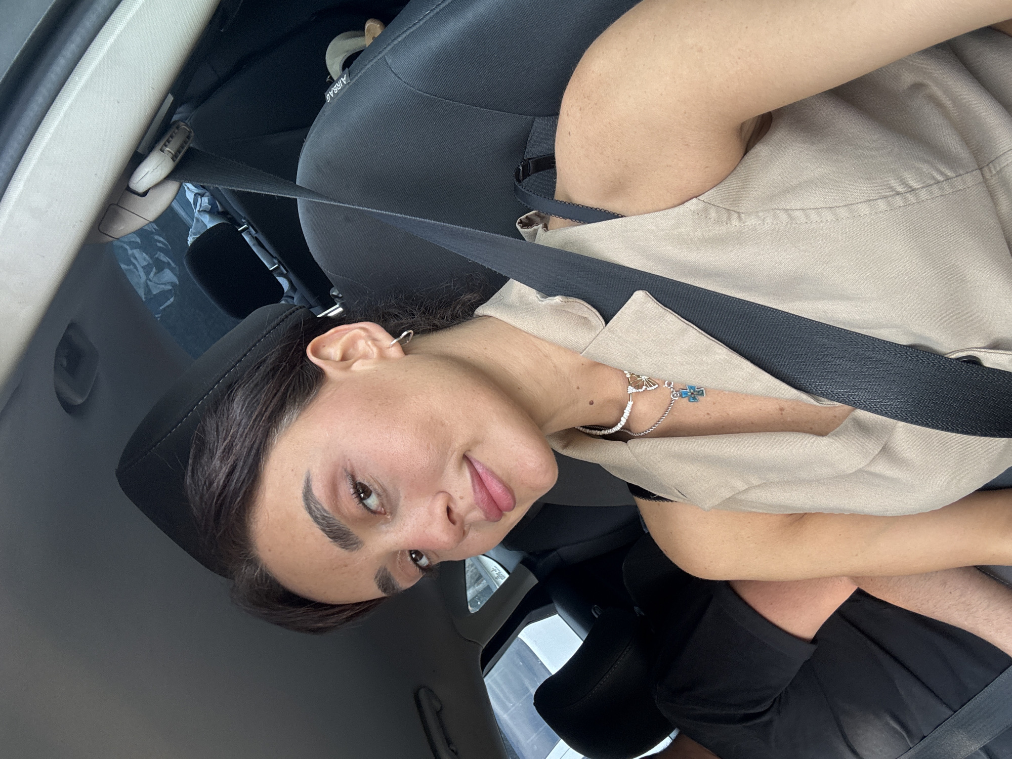 A young woman with dark hair and fair skin, wearing a beige sleeveless vest and a necklace with a blue cross pendant, is taking a selfie in the passenger seat of a car. A seatbelt is visible across her chest. To her left, a person wearing a black t-shirt is visible. The interior of the car, including the headrest and seatbelt mechanism, is also visible. The image appears to have been taken during the day. The location of Tbilisi, Georgia, is not discernable from the image itself.