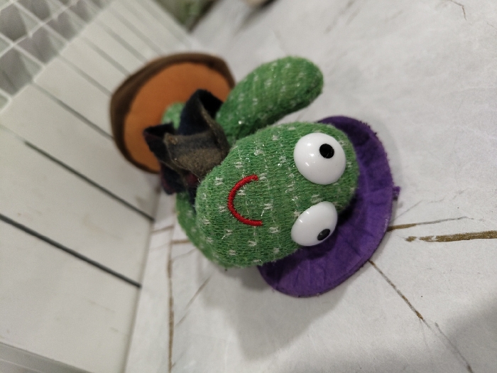A green plush toy with white, googly eyes, a red smile, and a purple felt base sits on a white, patterned surface. It wears a brown hat with a black brim, reminiscent of a mushroom. The background includes a white, slated structure. The toy's textured green fabric suggests a handmade quality.