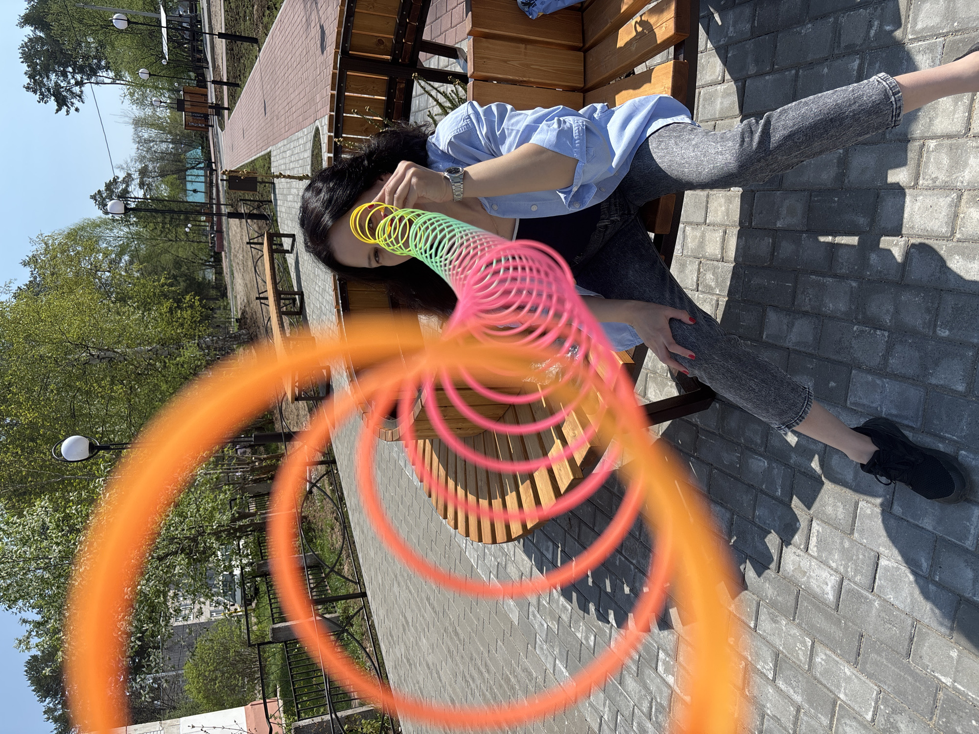 A woman with long, dark hair is sitting on a wooden bench on a paved surface in what appears to be a park in Chaikovsky, Russia. She is wearing a light blue button-up shirt, a dark top underneath, and gray jeans. She has a watch on her left wrist and is holding a colorful slinky toy near her face. The slinky is stretched out in a spiral shape, with colors ranging from orange to yellow to green to pink.
The background includes trees, street lamps, and other benches. The paved surface is made of gray bricks. The picture's orientation makes it appear as though the image is tilted.