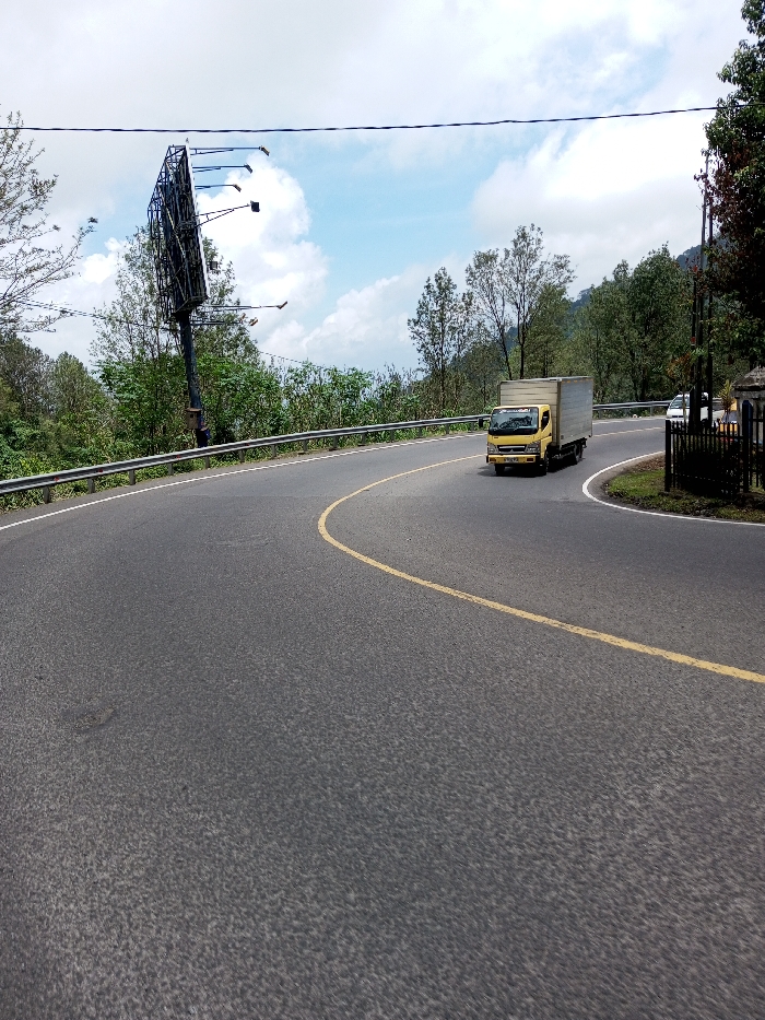 The scene depicts a curved asphalt road in the city of Boommlog, Bhutan, under a partly cloudy sky. A yellow box truck with a white cargo container proceeds along the right side of the road, navigating a rightward curve. The road surface is dark gray asphalt, marked with a continuous yellow line on its inner arc and a solid white line on its outer edge. A silver metal guardrail runs along the left side of the road. To the left of the road, an empty black metal billboard frame, featuring several outward-projecting light fixtures, stands amidst dense green trees and undergrowth. On the right side, a paved turn-off branches from the main road, bordered by a black metal fence. Two white vehicles are partially visible behind this fence. Overhead utility lines are present across the upper frame. The surrounding environment consists of lush, tree-covered hills. No individuals are visible.