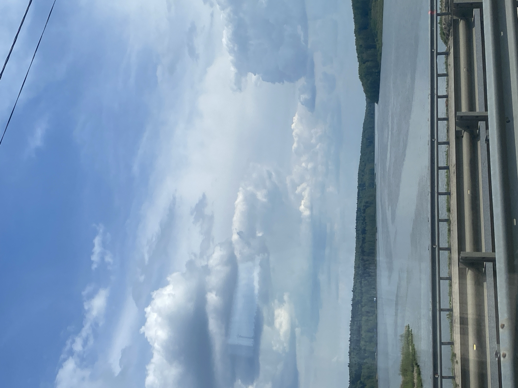The image shows a view from a bridge over a body of water. The sky is predominantly blue with scattered cumulus clouds. Along the right edge of the image, there is a bridge railing and support structure. Beyond the railing is the body of water, with a wooded shoreline visible on the far side. A power line is visible in the upper-left corner.
Without further information, it is impossible to confirm if this image is located in Bereznyaki, Russia.