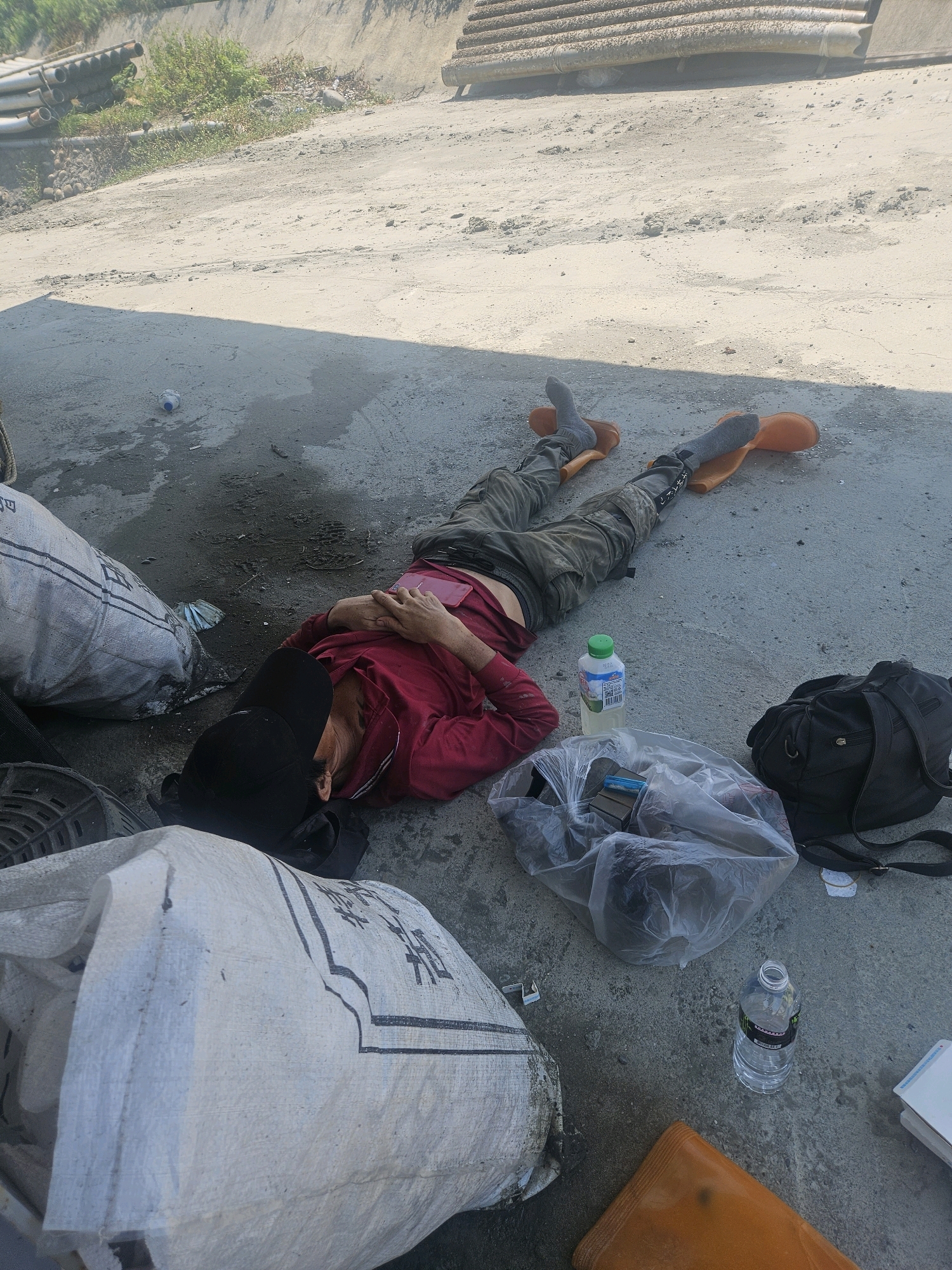 A person is lying on their back, asleep, on a concrete surface. They are wearing a red long-sleeved shirt, dark pants, grey socks, and bright orange boots. A black baseball cap is pulled down over their forehead, and their hands are clasped over their chest. A pink object, possibly a phone or wallet, is resting on their chest.
Surrounding the person are several objects. To their left is a large white woven bag with black lettering. To their right is a black backpack, a clear plastic bag containing items, and a bottle of clear liquid with a green cap. Another water bottle is visible further to the right.
The setting appears to be an outdoor, possibly industrial or construction area, given the concrete ground, the presence of large bags that could contain materials, and some pipes visible in the background. The lighting suggests it is daytime, with shadows indicating the sun is out. The overall scene conveys a sense of rest or exhaustion. The white bag has Chinese characters, but they are not clearly legible. The liquid bottle has some text and a green cap.