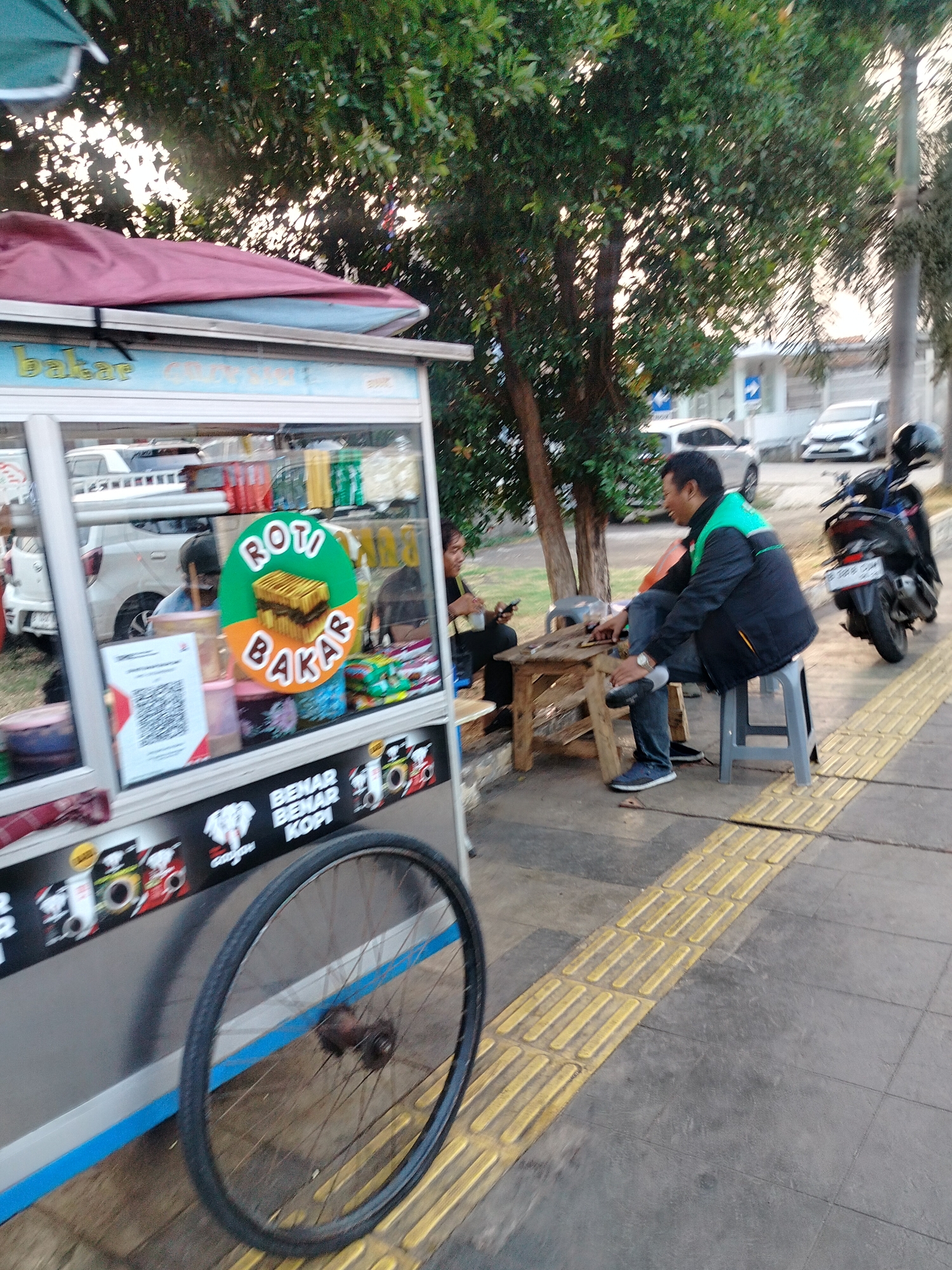 The image depicts an outdoor scene on a city sidewalk in East Jakarta, Indonesia. In the foreground, a wheeled food cart selling "Roti Bakar" (grilled bread) is prominent, adorned with graphics and signage. A bicycle wheel is attached to the side of the cart. To the right of the cart, a man wearing a black and green jacket and blue jeans is seated on a grey stool, looking towards the cart. He is holding an object, possibly a phone. Another person is partially visible in the background, seated on a low stool. The sidewalk has tactile paving in yellow. A motorcycle is parked to the right of the seated man. Several cars are parked or driving on the street in the background. Trees and vegetation are present, with a building visible behind them. The lighting suggests it is daytime, possibly late afternoon or early morning due to the angle of the sun.