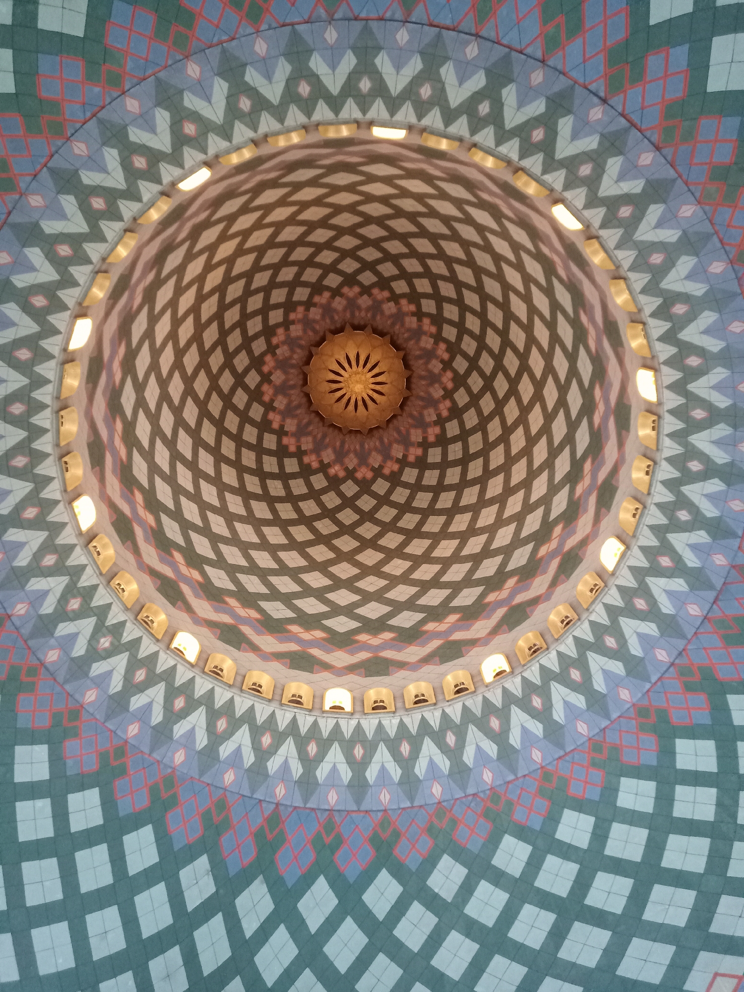 This is an upward-facing shot of the interior of a domed ceiling, likely at the Masjid Al-Akbar in Surabaya, Indonesia. The dome features intricate geometric patterns in shades of teal, blue, pink, and white. The outer portion of the dome displays a repeating pattern of diamonds and arrow-like shapes. Closer to the center, the pattern transitions to a grid of diamonds, with diagonal lines creating a lattice effect. Along the rim where the patterned dome meets the central section, there is a series of arched windows, each containing a warm, illuminated light fixture. At the apex of the dome, a decorative, starburst-shaped golden fixture hangs, casting subtle shadows. The lighting creates a warm and inviting atmosphere.