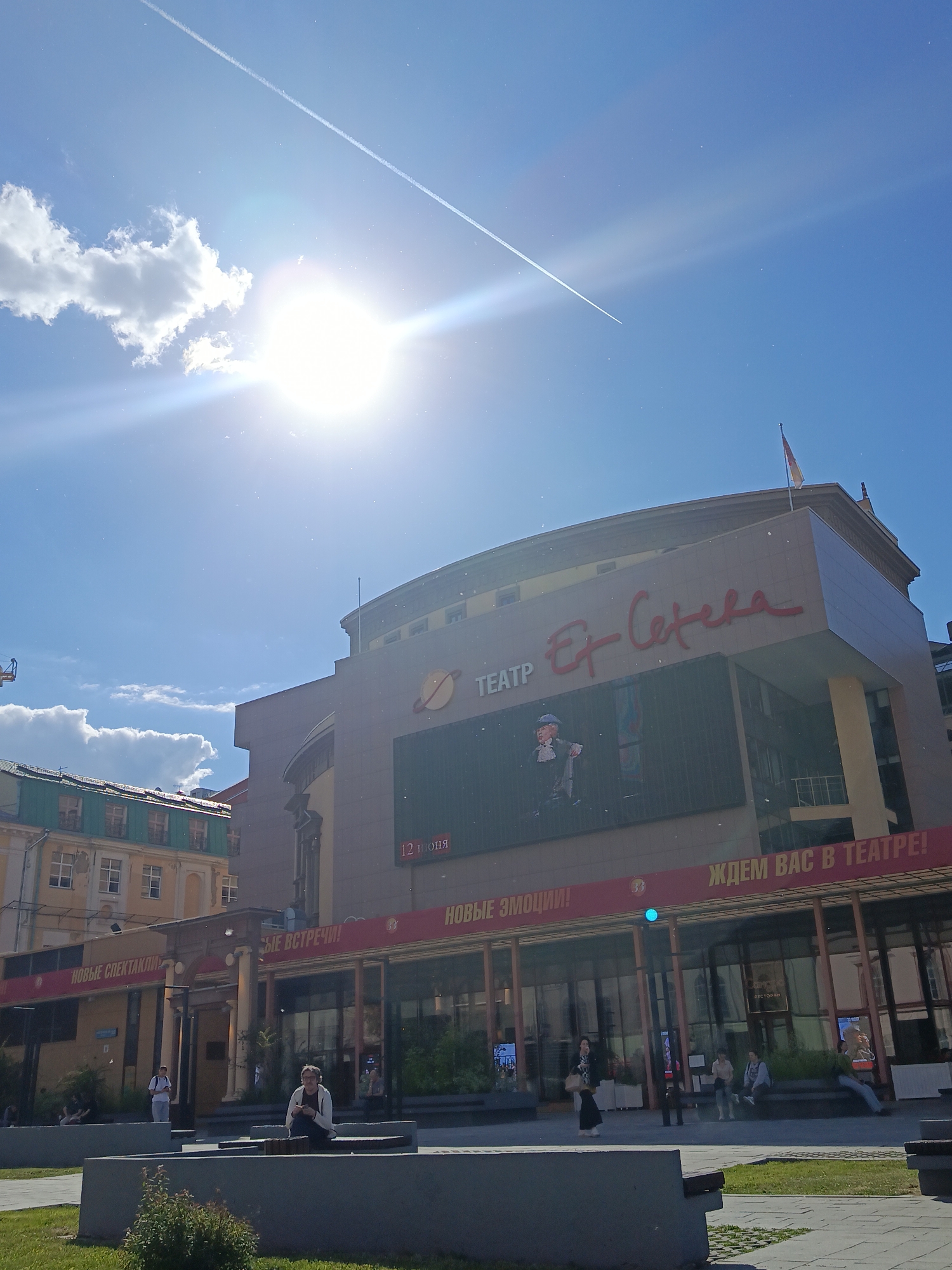 The image captures an exterior view of the "Et Cetera" Theater on a bright, sunny day. The large, modern building dominates the center-right of the frame, featuring a tan facade with a prominent red "ТЕАТР Et Cetera" sign. Below the main sign, a large LED screen displays an image of a character (possibly from a play) and the text "12 ІЮЛЯ" (12 July).
The ground floor of the theater has a glass facade with banners above it. Visible text on these banners includes "НОВЫЕ СПЕКТАКЛИ" (New performances), "НОВЫЕ ВСТРЕЧИ!" (New meetings!), "НОВЫЕ ЭМОЦИИ!" (New emotions!), and "ЖДЕМ ВАС В ТЕАТРЕ!" (We are waiting for you at the theater!).
In the foreground, there's an urban public space with concrete benches and a small patch of grass. Several people are visible in this area: one person is seated prominently on a bench in the lower middle, facing left, while others are seated or standing further back near the theater entrance. They appear to be casually waiting or relaxing.
The sky is a clear, vibrant blue with some scattered white clouds. The sun is intensely bright and directly facing the camera, creating a strong lens flare and sunburst effect in the upper left quadrant. A distinct white contrail from an airplane stretches diagonally across the sky from the upper left to the upper right. Fine dust particles or specks are visible throughout the sky, illuminated by the strong sunlight. To the left of the theater, an older building with a green roof and multiple windows can be seen. The overall impression is a lively, sunlit urban scene.