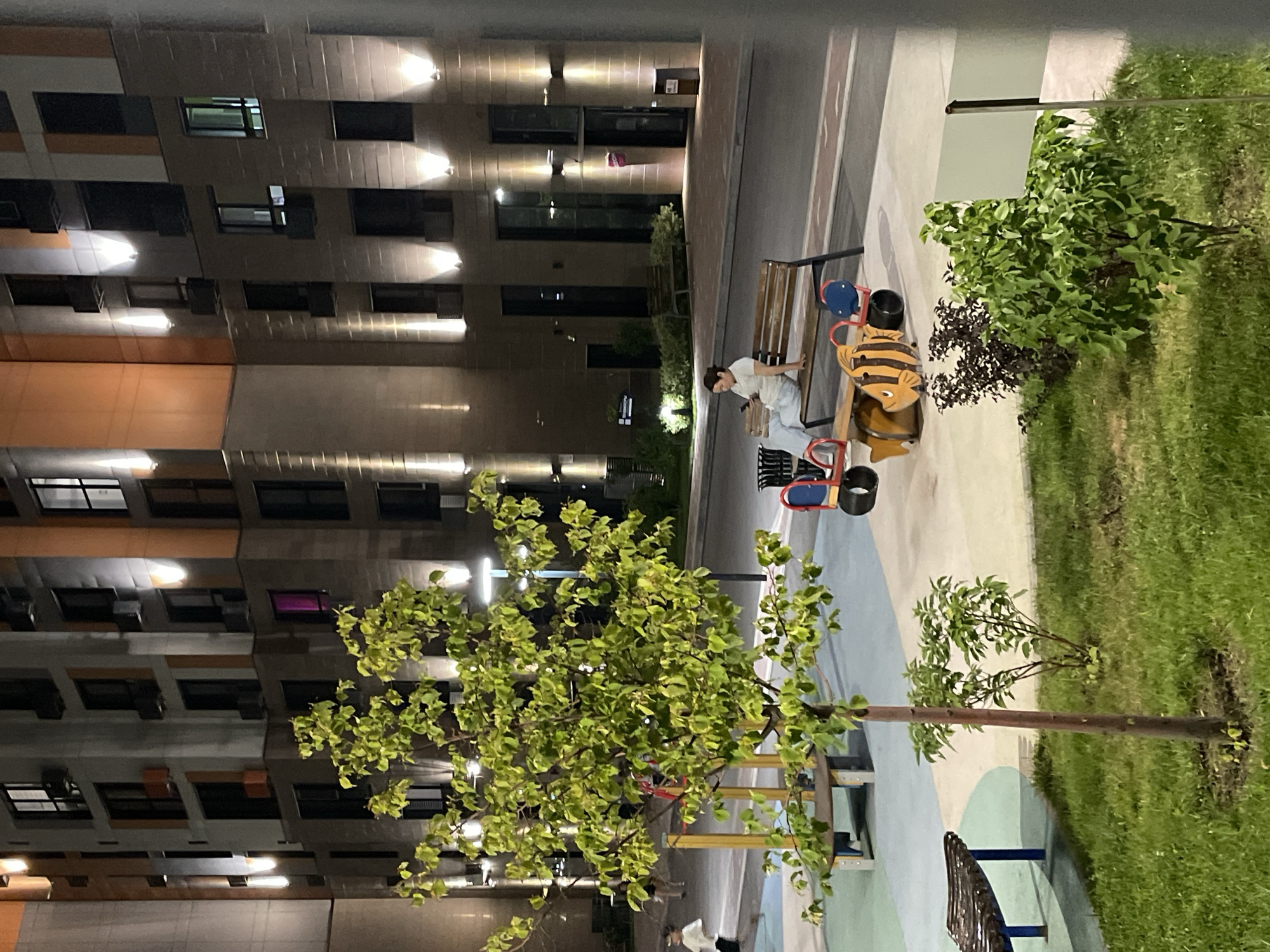 This is a nighttime exterior photograph of a modern residential complex, likely in the city of Almaty, Kazakhstan. The foreground features a small tree with vibrant green leaves and a patch of grass. To the left, a section of a playground with a wooden bench is visible. In the middle ground, a woman sits on a park bench facing right, looking at her phone. Adjacent to the bench is a whimsical playground ride shaped like a bee. Further back, more playground equipment and what appears to be a trash can are present. The background is dominated by the illuminated facade of a multi-story apartment building, characterized by its grid of windows and architectural lighting. The building's exterior displays a mix of light and dark materials, with warm orange accents. The overall impression is one of a well-lit, contemporary urban residential area after dark.