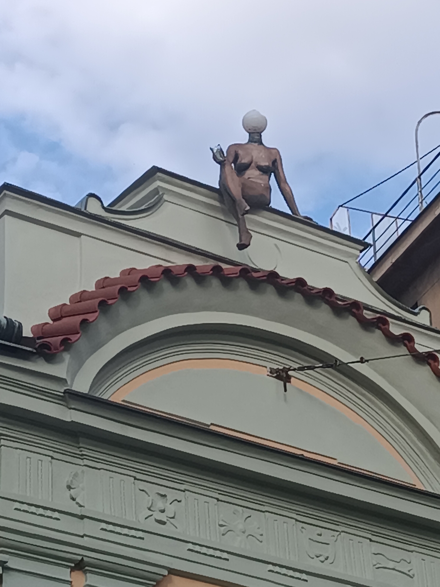 In Prague, Czechia, a bronze sculpture of a nude woman is perched atop a building. The sculpture depicts a woman sitting with her legs crossed and holding a glass in her right hand. Her head is a glowing orb, and her body is rendered in a realistic style. The sculpture is situated on a roof adorned with red tiles, and behind it, a blue sky with scattered clouds can be seen. The building itself features classical architectural elements, including ornate detailing and arched structures. The overall scene captures a unique piece of public art integrated into the urban landscape of Prague.