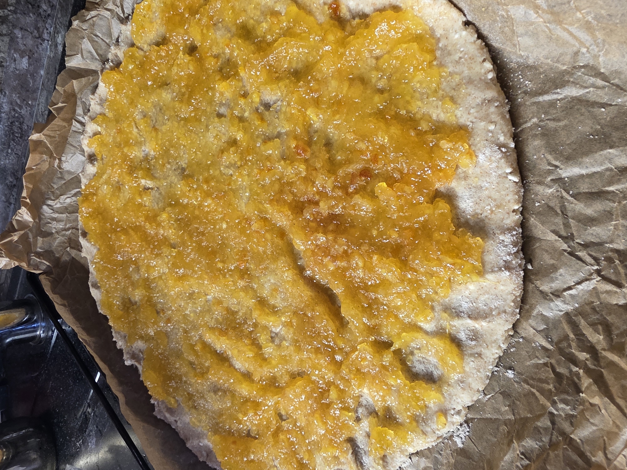 The image shows a close-up, overhead view of a rustic, whole-wheat pizza base that has been spread with a thick, vibrant orange, possibly fruit-based, topping. The topping has a slightly lumpy, jam-like texture with visible pieces of fruit or zest, and it appears to be sprinkled with fine sugar crystals, giving it a glistening sheen. The pizza base is circular and unevenly shaped, resting on crinkled brown parchment paper, which in turn is placed on a dark countertop. The edge of the pizza base is dusted with white powder, likely flour. In the background, a stainless steel kitchen faucet is visible, along with a glimpse of a dark, reflective surface, suggesting a kitchen environment. There are no people or animals present in the image. The scene does not provide any information that would allow for identification of its specific location within Saint Petersburg, Russia.